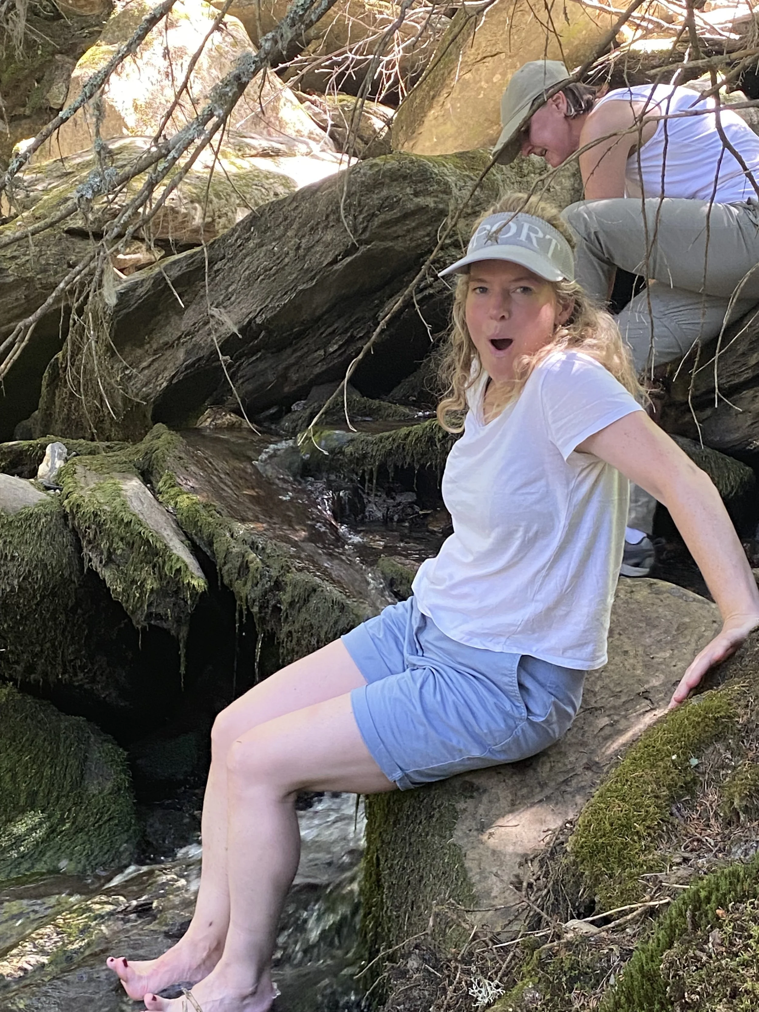 Two women on rocks in a creek, one sitting with her feet in the water and the other kneeling behind her, surrounded by mossy rocks and overhanging branches in a forest.