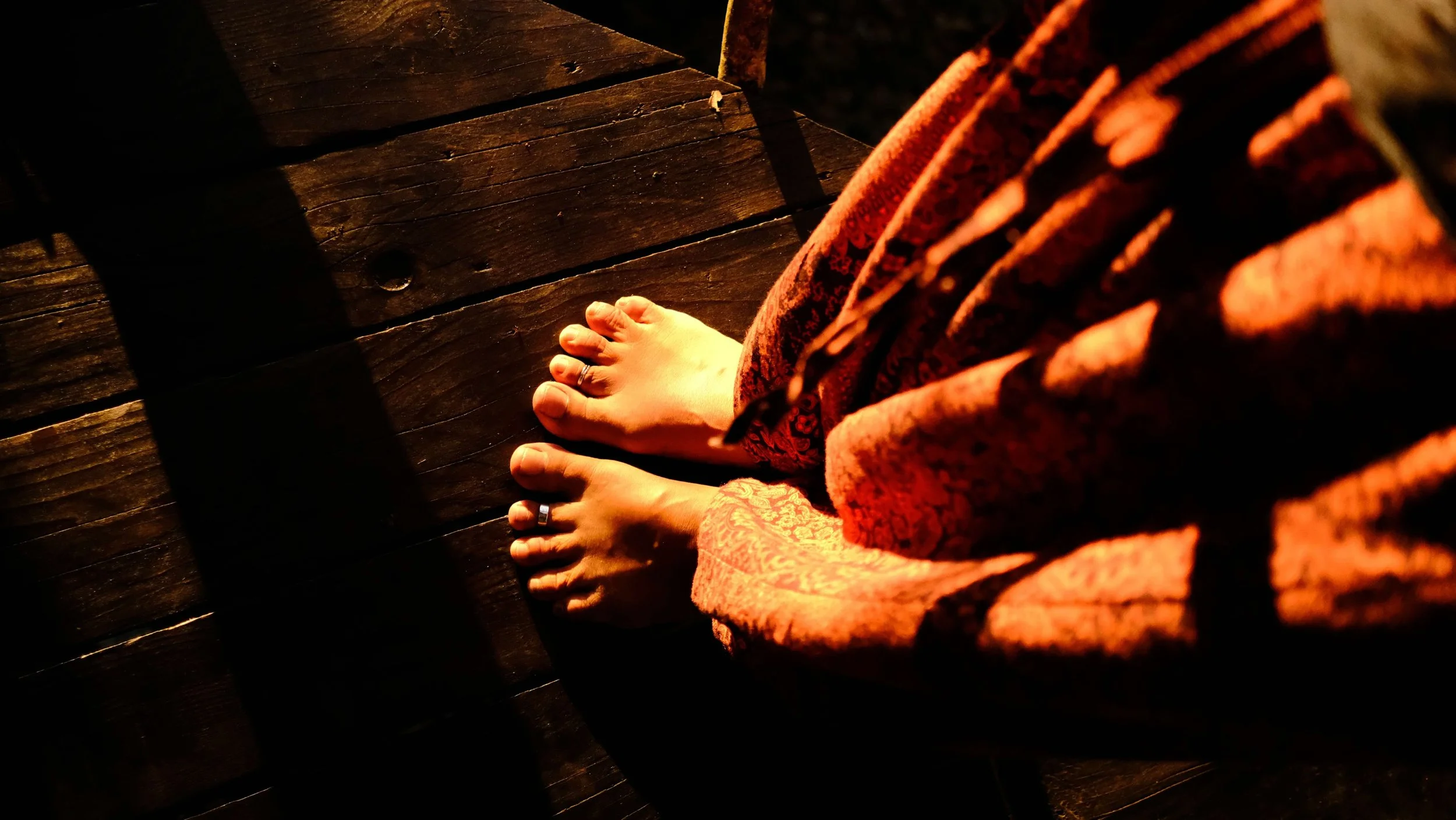 Person standing on wooden floor, wearing a patterned red garment, with bare feet and wearing rings on toes and fingers.