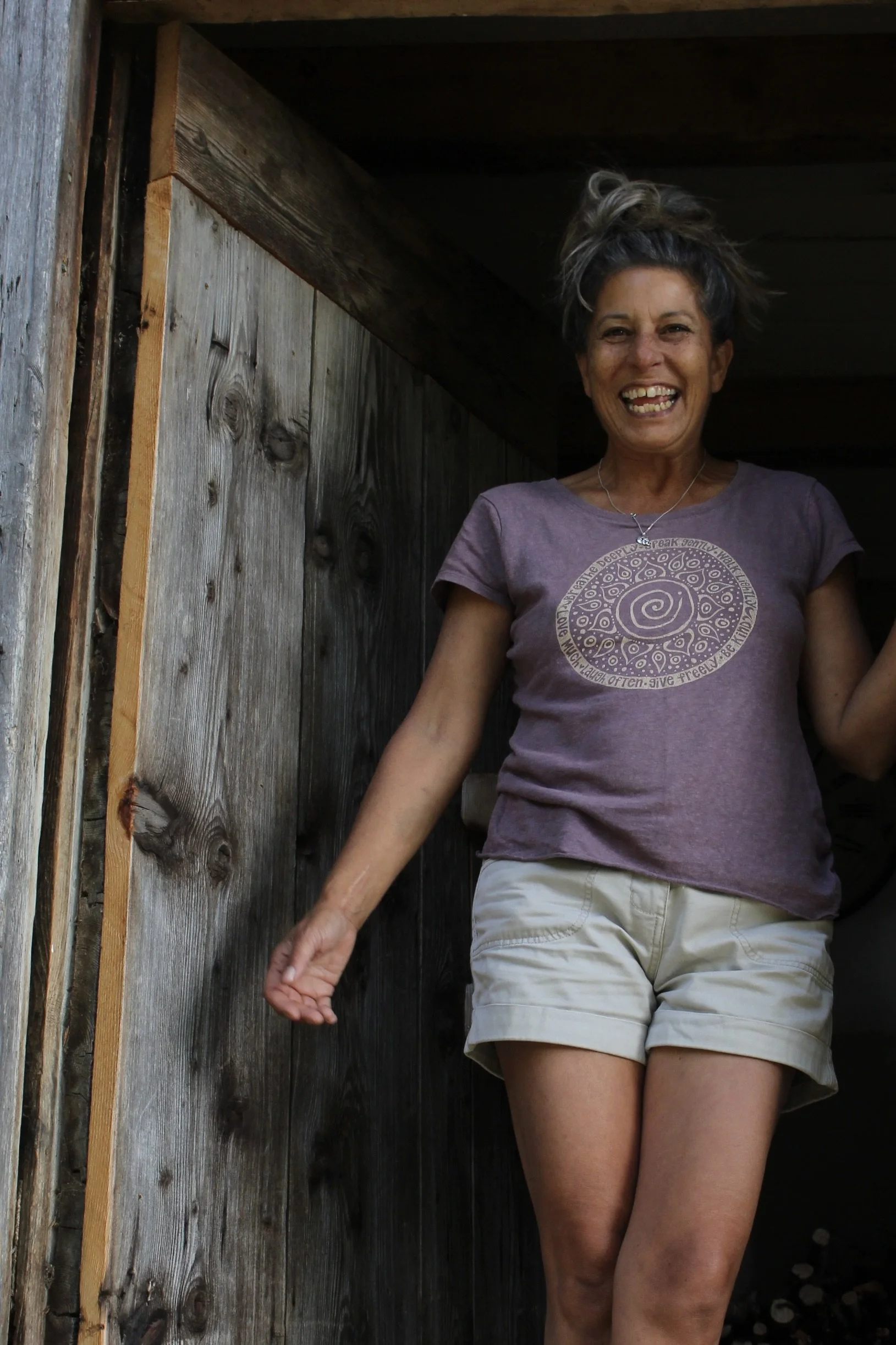 Soraya smiling in doorway of cabin with gray hair hair tied up, wearing a purple t-shirt