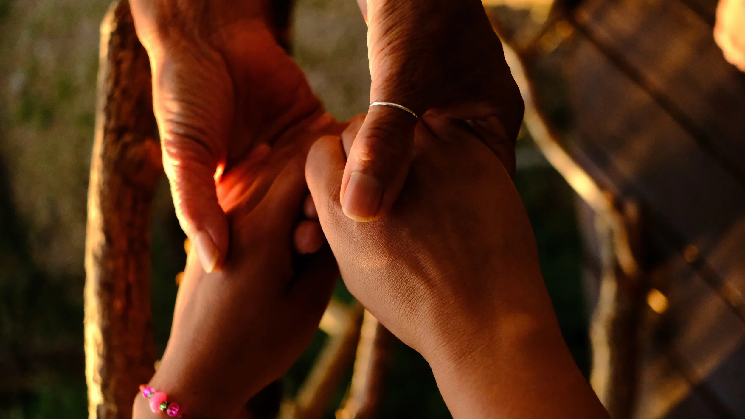 Close-up of two hands holding each other, with one hand wearing a silver ring and a colorful beaded bracelet, against a blurred background of tree branches.