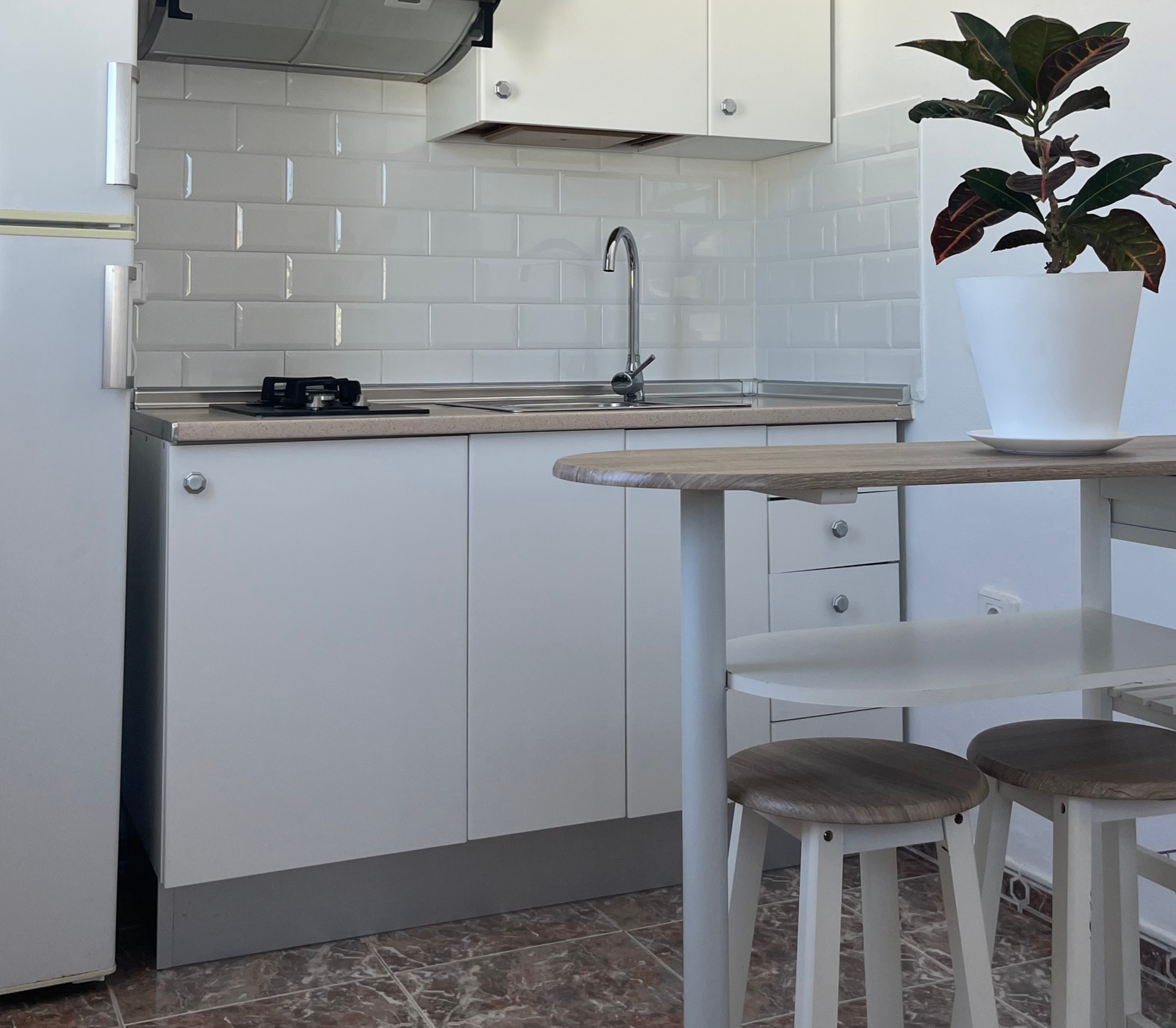 Small kitchen with white cabinets, a refrigerator, microwave, stove, sink, and a small table with two stools and a potted plant.