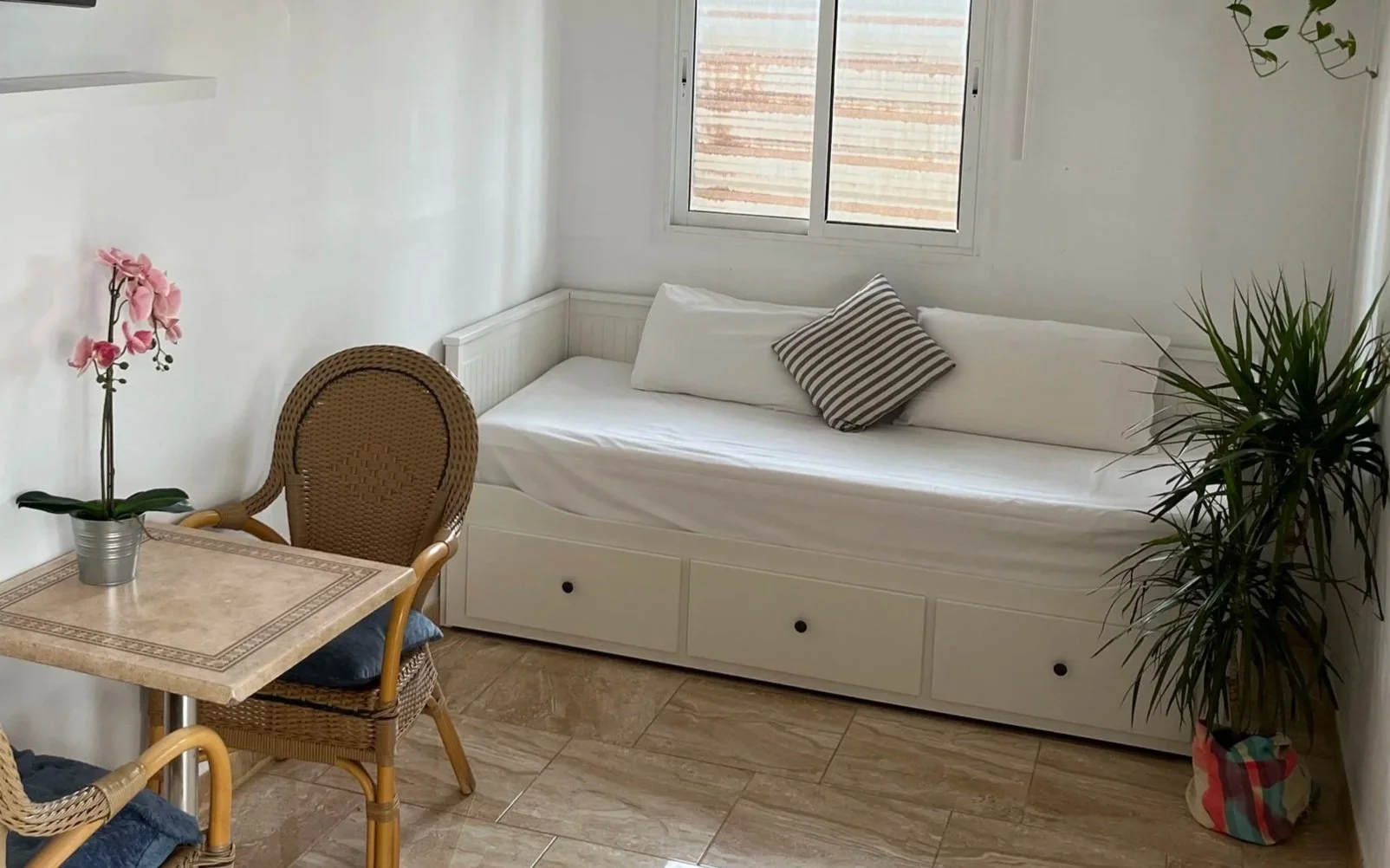 Small living room with white walls and tiled floor, featuring a wall-mounted TV, a window with gray blinds, a white daybed with a striped pillow, a small table with two wicker chairs, a pink orchid plant, and several potted plants.