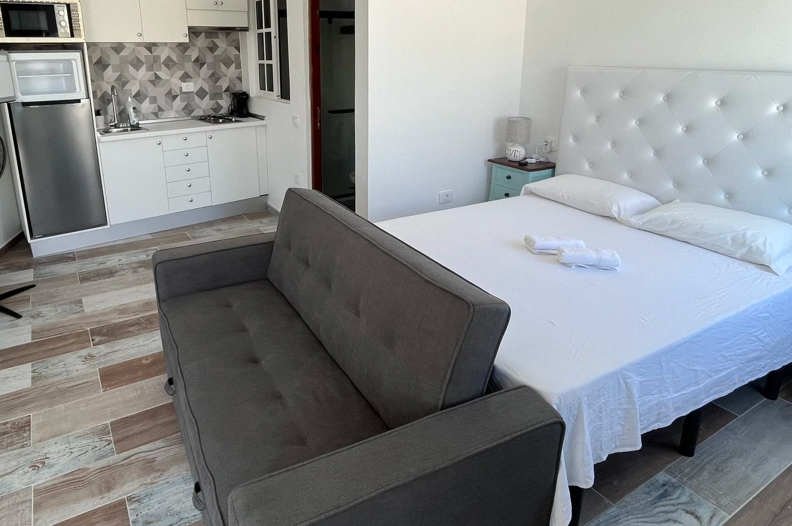 Studio apartment with a bed, a sofa, a kitchenette, and a small dining table, featuring white walls and wooden flooring.