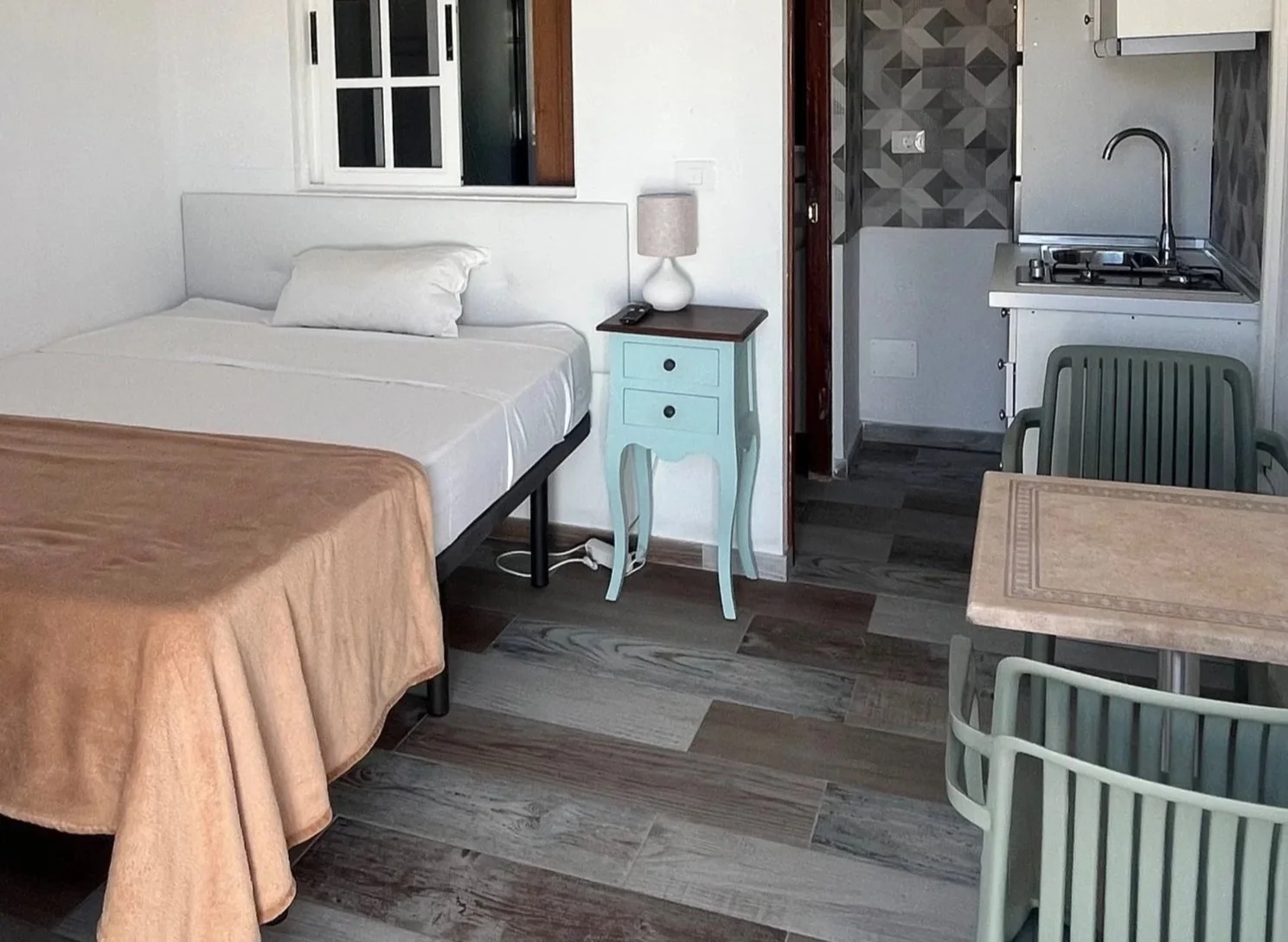 A room with a bed, small nightstand with a lamp, small table with two chairs, kitchenette with sink and stove, and a window with white frame.
