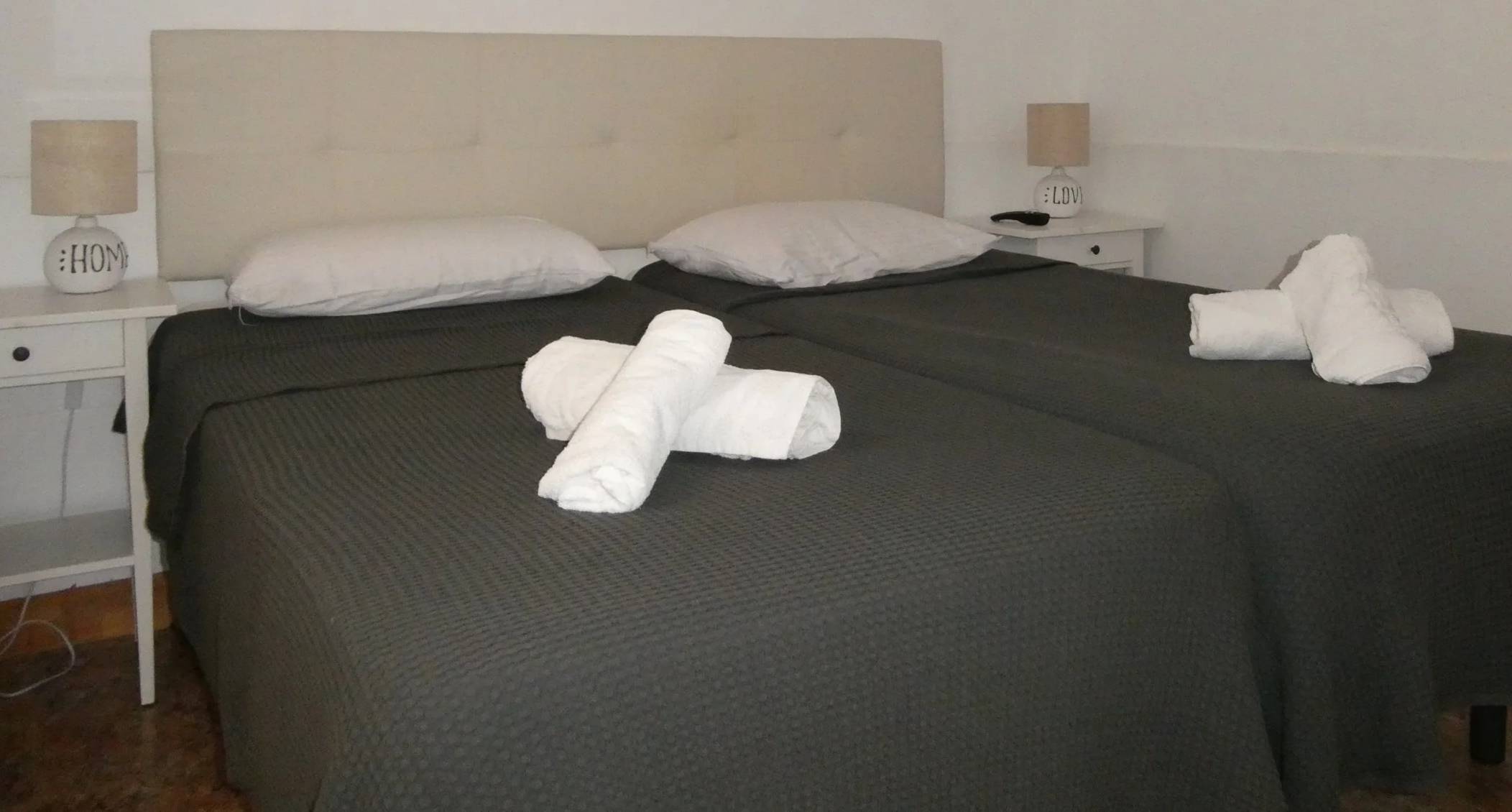 A hotel room with two single beds covered in dark gray bedspreads. Each bed has a white pillow and towel art shaped like a dog or bear. There are white nightstands on each side of the beds with lamps, and the walls are plain white.