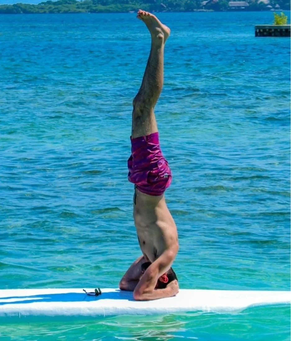 Man balancing in a headstand on a paddleboard in clear blue water, wearing purple shorts, with a distant shoreline in the background.