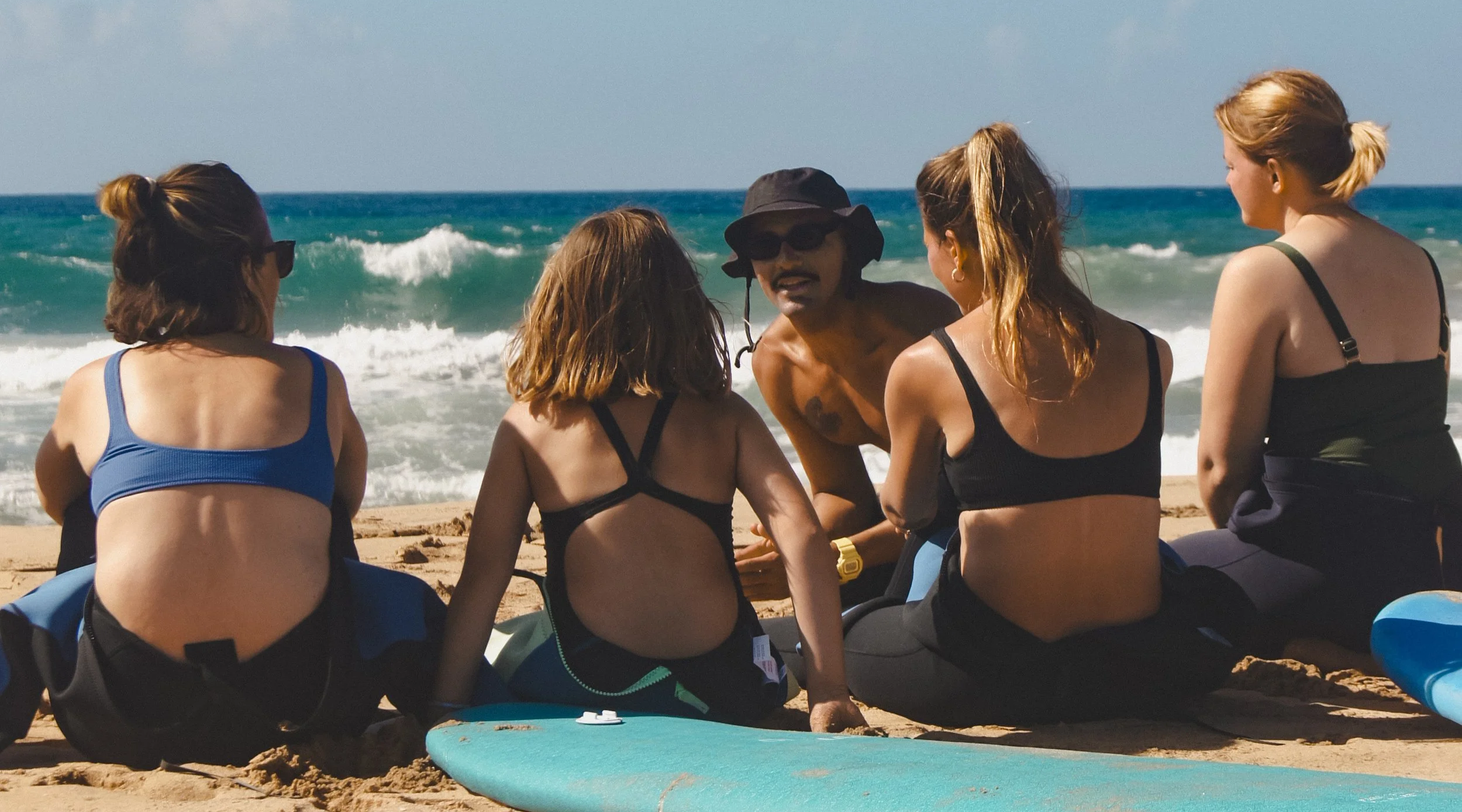 A group of five people, including a man with a hat, sitting on the sandy beach near the ocean, conversing with each other amidst surfboards.