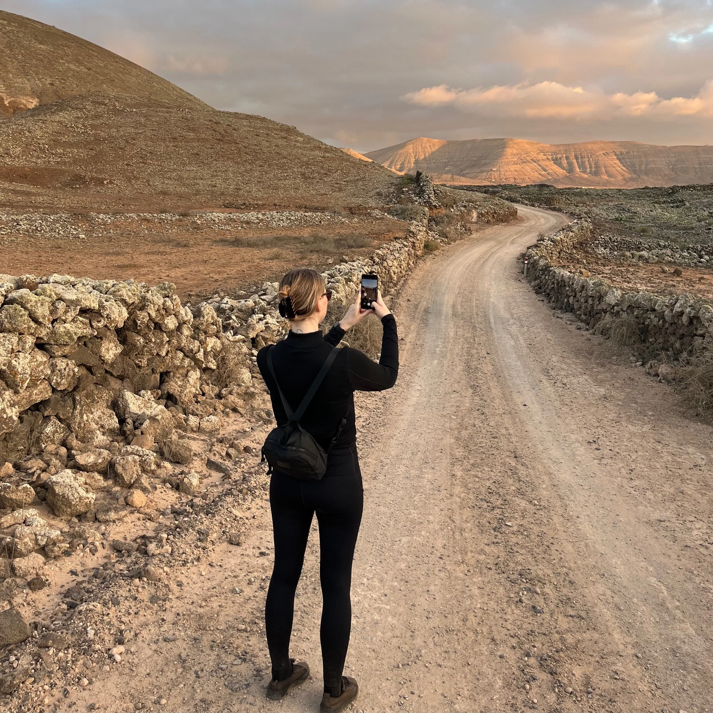 A woman in black clothing taking a photo of a winding dirt road through a desert landscape with rocky hills and mountains in the background.