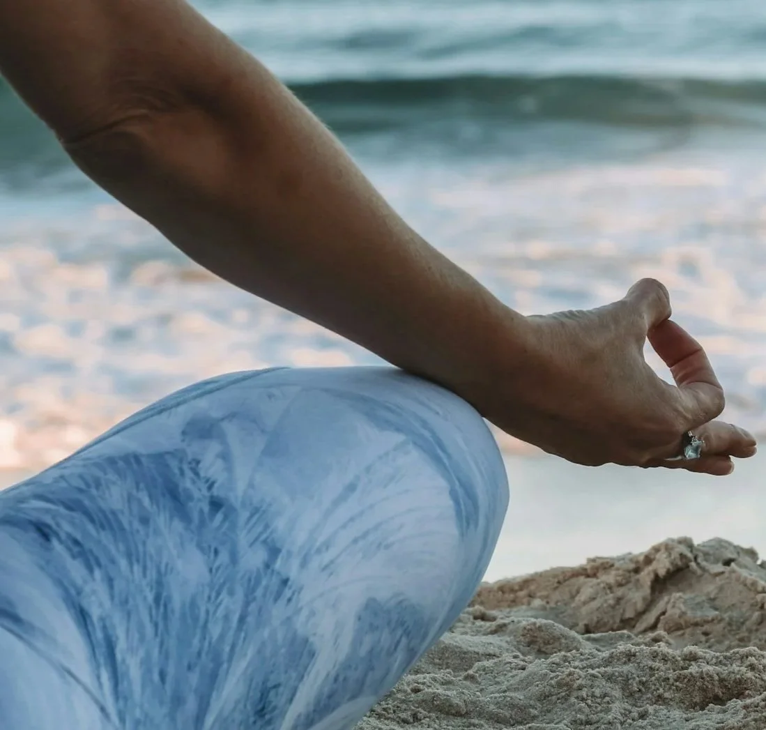 Person sitting on the sandy beach in a cross legged yoga pose, making a mudra with their hand, near the ocean with gentle waves in the background.