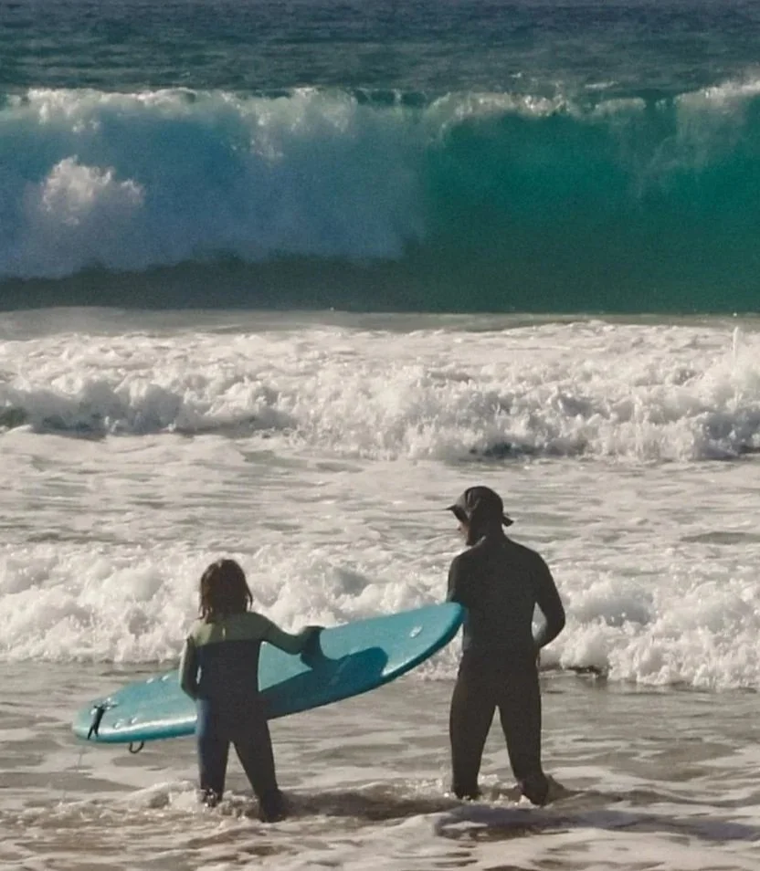 A person and a child holding a surfboard stand in shallow water at the beach with large waves in the background.