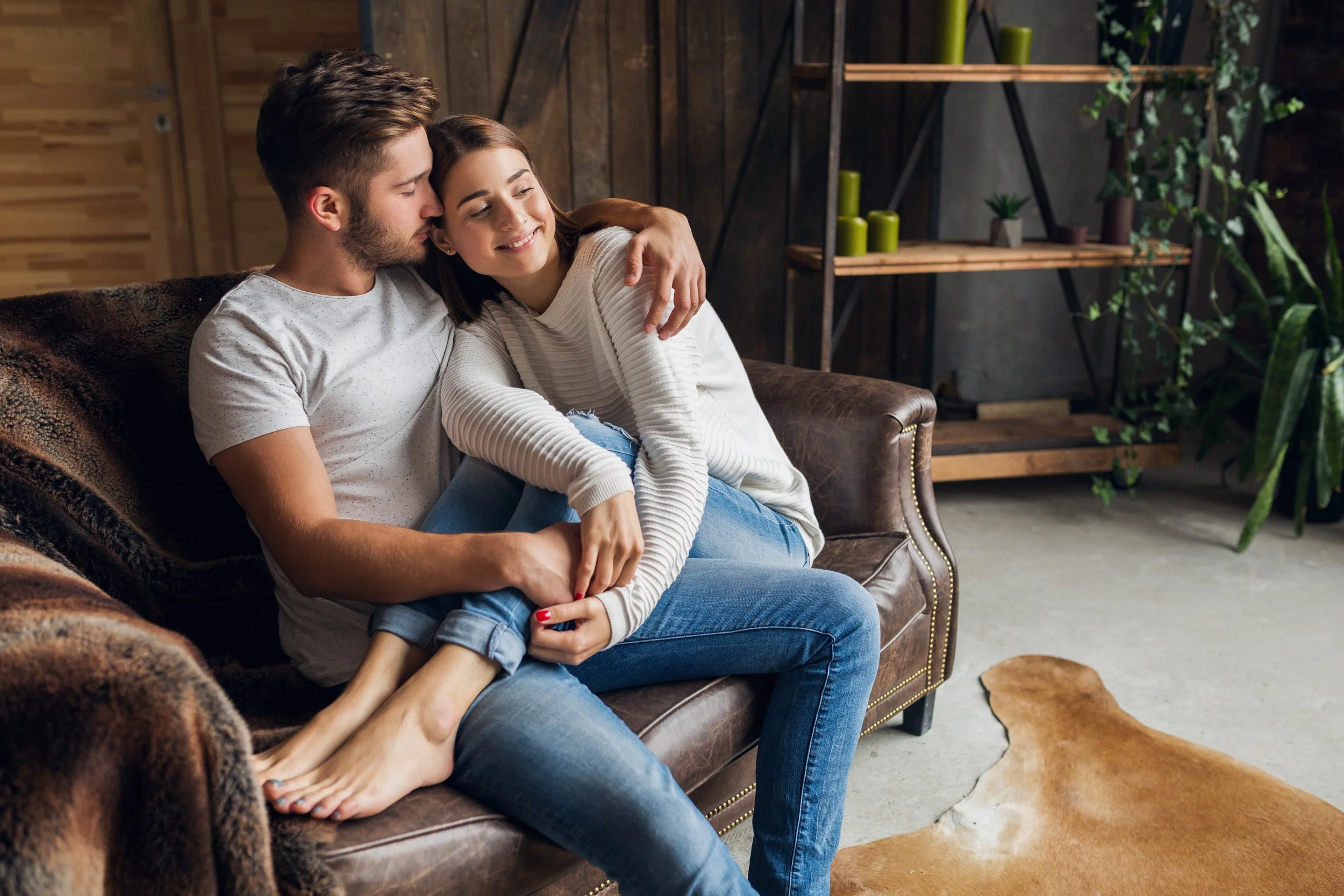 A young couple sits on a brown leather couch in a cozy living room, with the man wrapping his arms around the woman who is smiling with her eyes closed. The room features wooden walls, green plants, and a wooden shelf with decorative items.