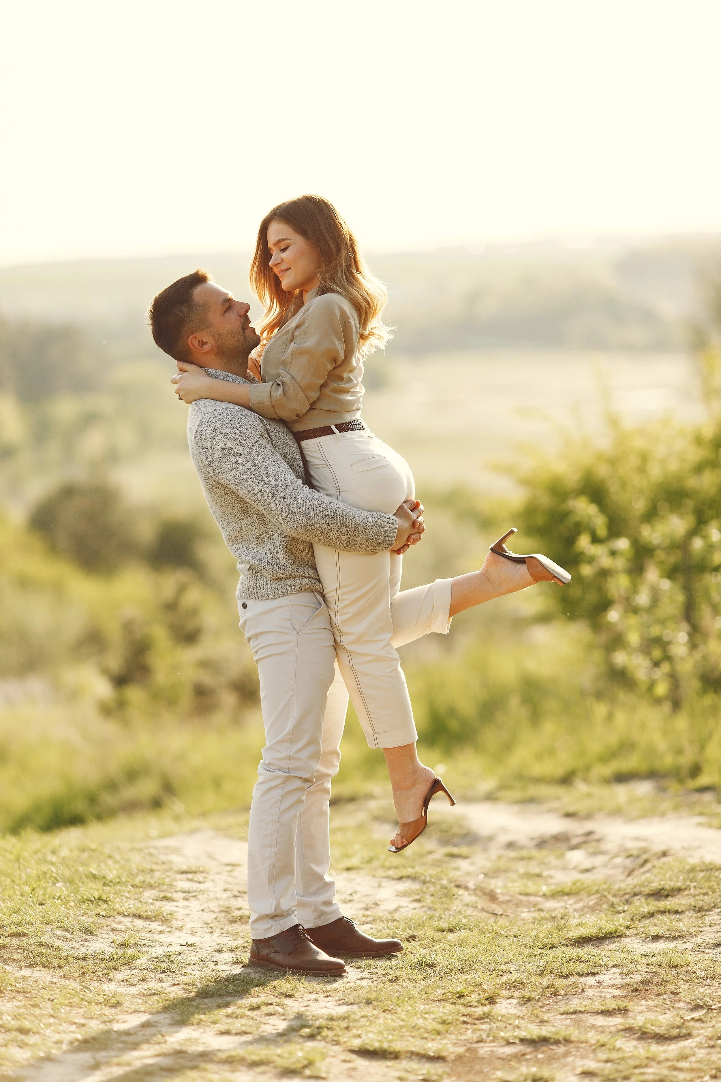 A man is holding a woman in his arms outdoors during sunset, with the woman smiling and looking at the man, as she is lifted off the ground in a grassy area.