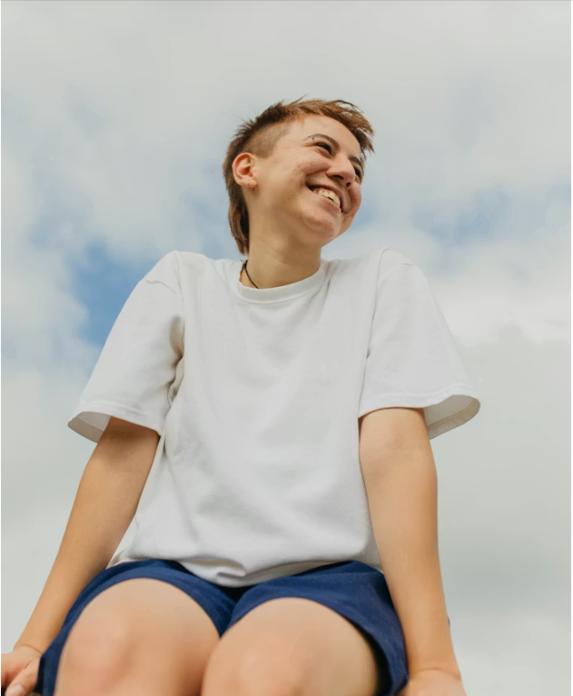 Young person with short hair smiling, wearing a white t-shirt and dark shorts, outdoors against a cloudy sky.
