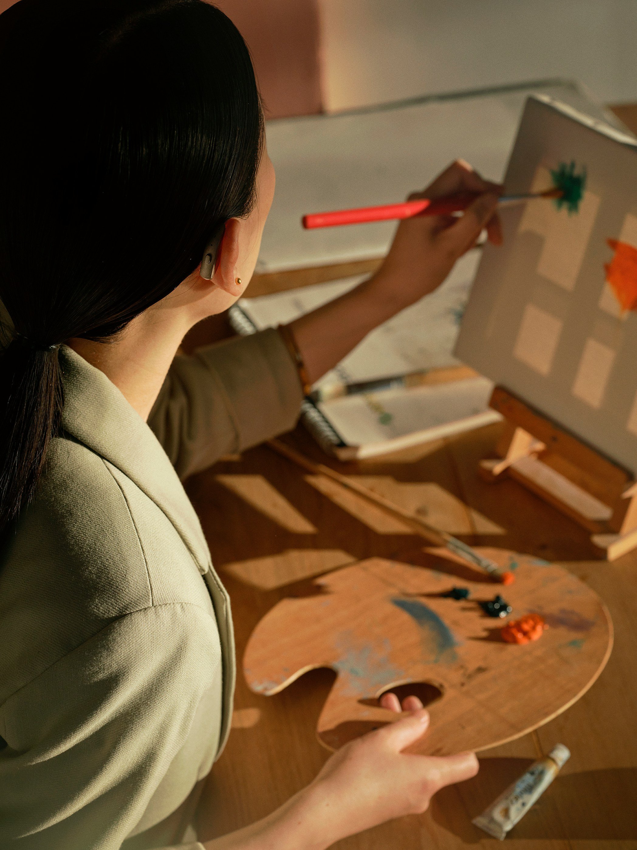 A person painting on a canvas with a small brush, standing at a wooden table with a painter's palette and tubes of paint.