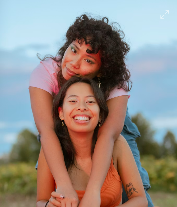 Two young women outdoors at sunset, one standing behind the other, both smiling and embracing each other.