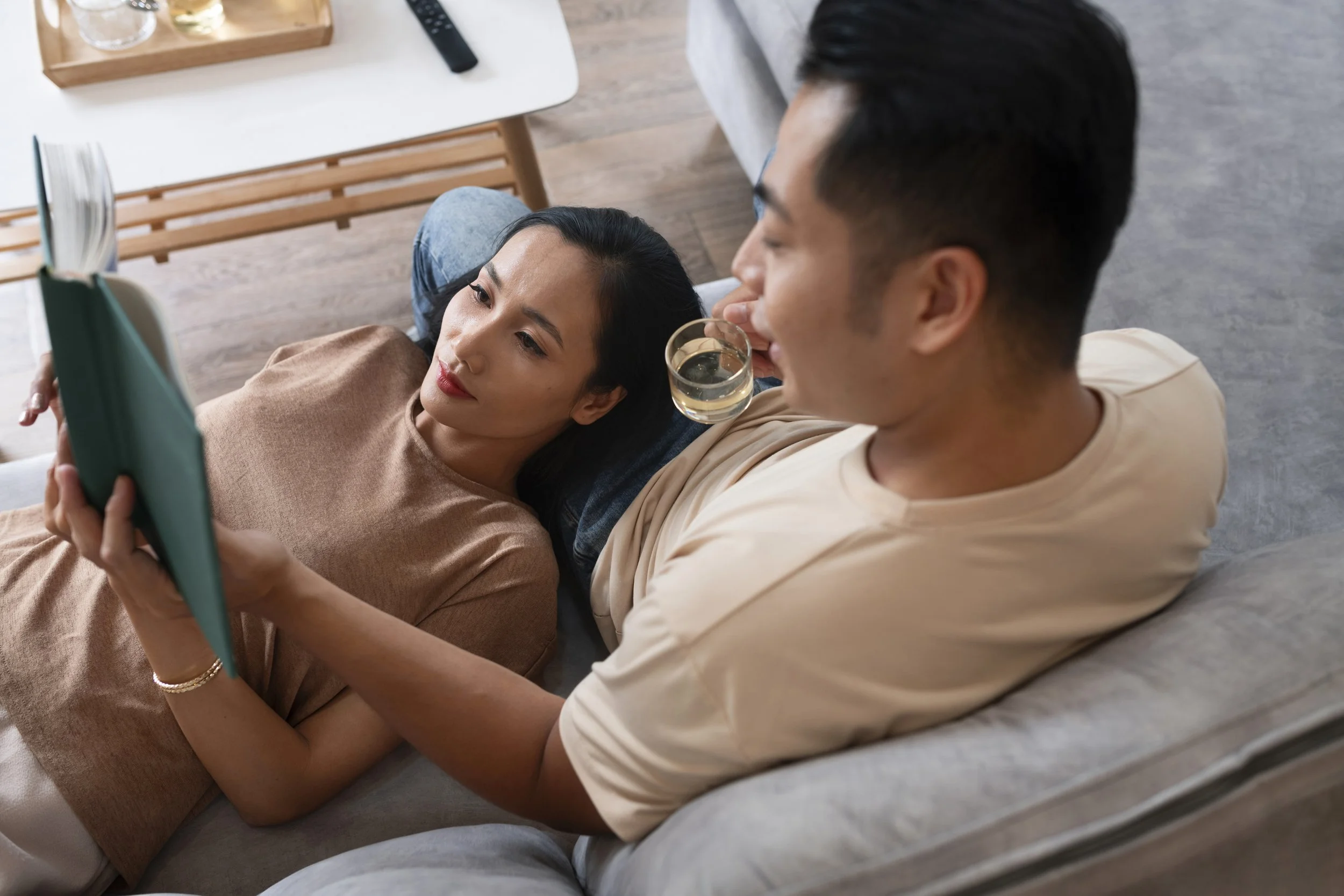 A couple relaxing on a couch, with the woman lying down and reading a book while the man holds a glass of wine and smiles at her.