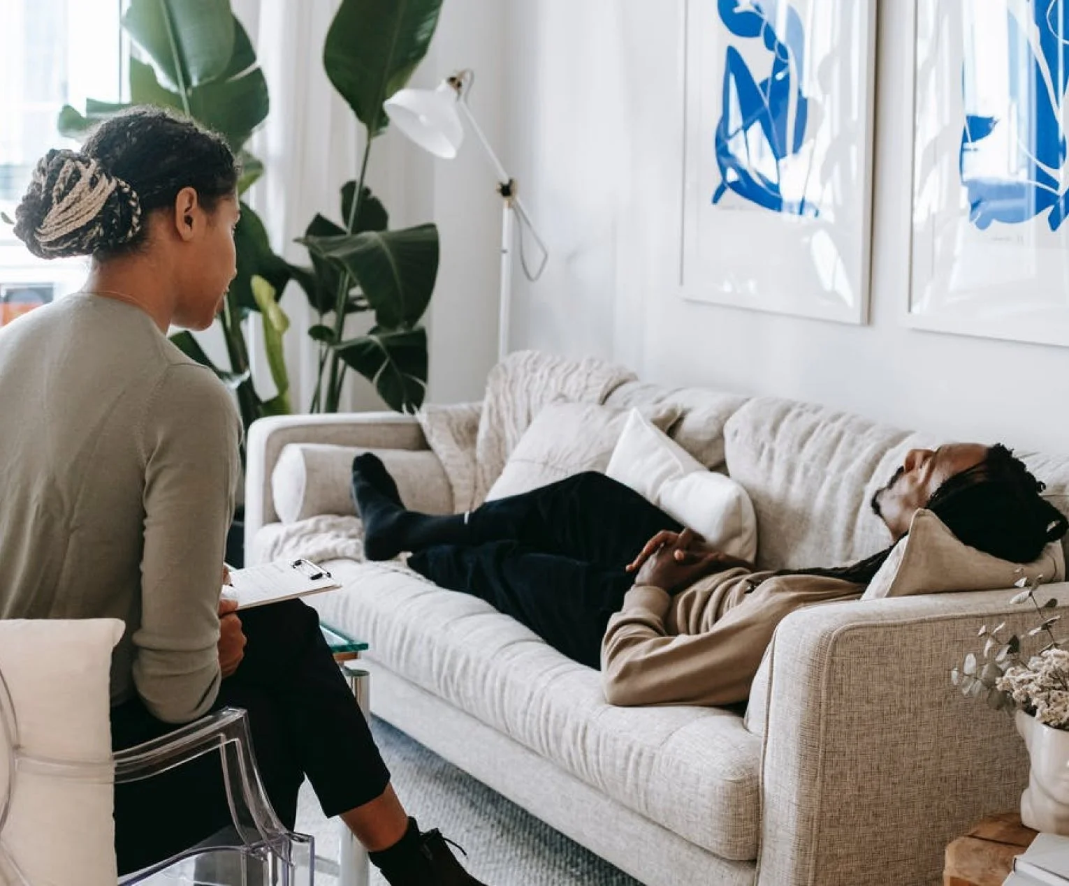 A woman lying on a beige couch and a woman sitting on a white chair having a conversation in a bright, modern living room with large green plants and blue abstract artwork on the wall.