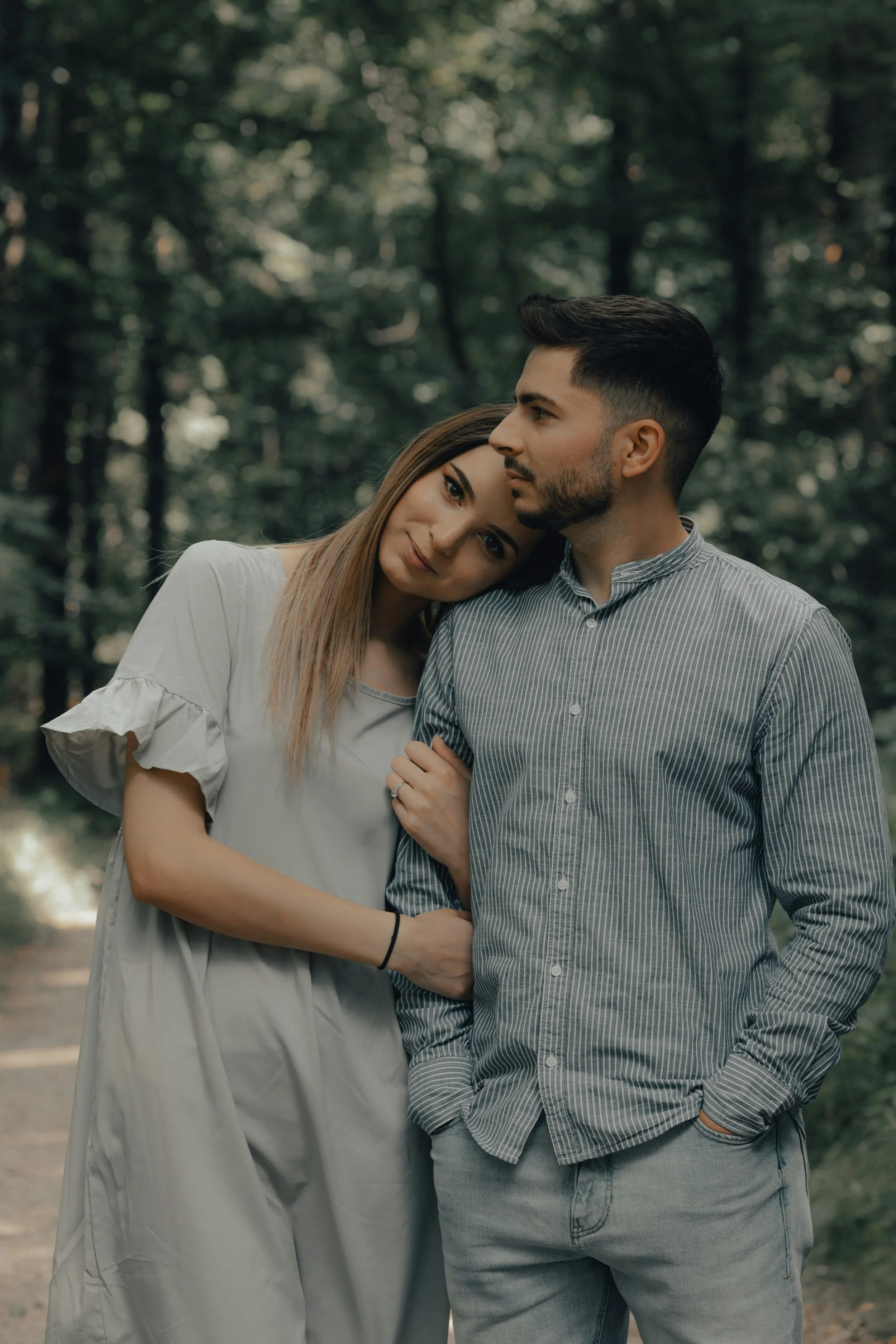 A young couple with light skin standing close in a wooded area, with the woman resting her head on the man's shoulder and both looking into the distance.