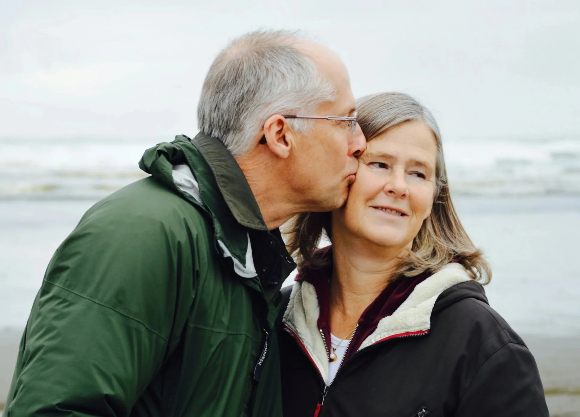 A middle-aged man is kissing a woman on the side of her head while they stand close together outdoors near the beach.