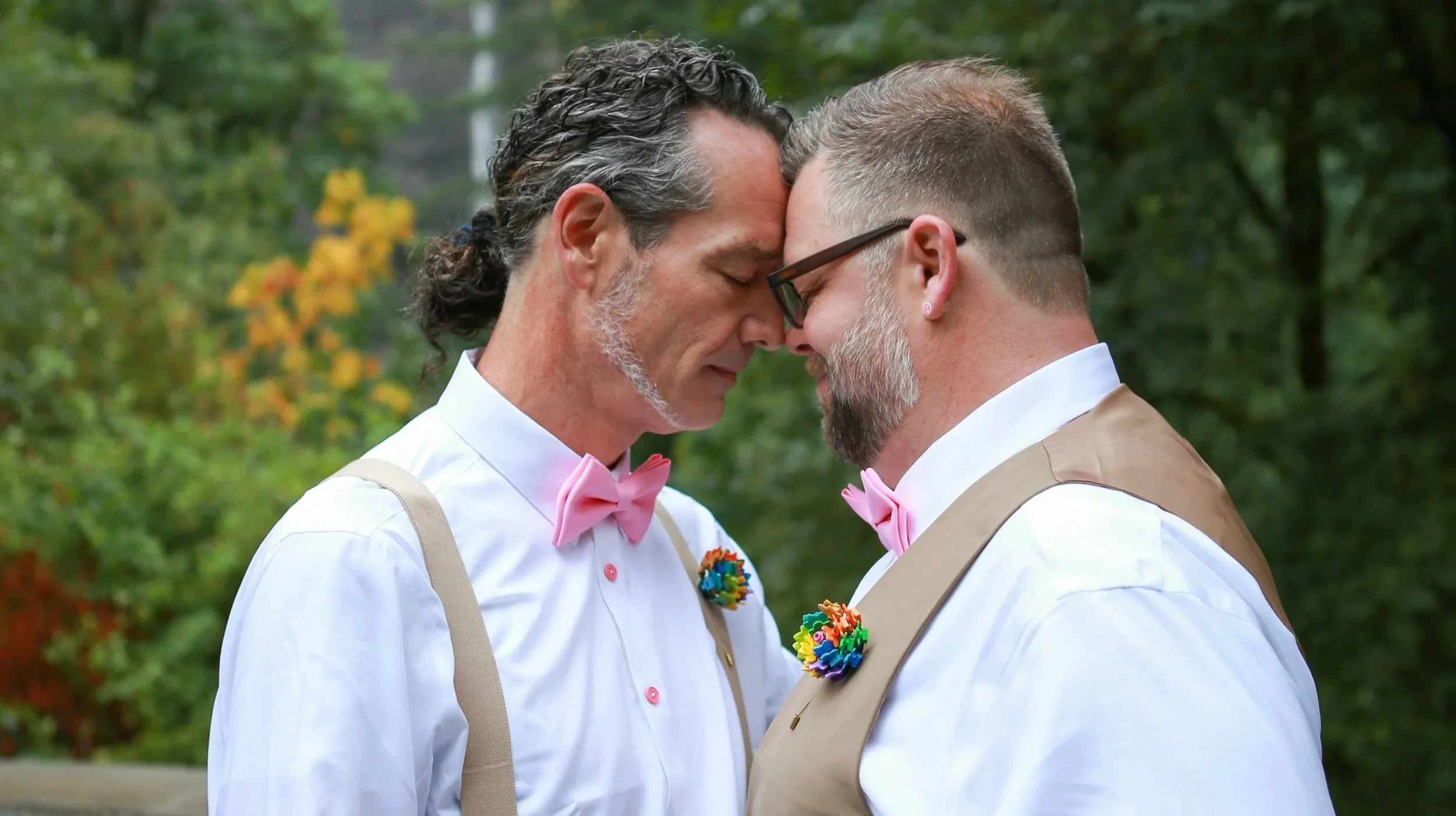 Two men with their foreheads touching, dressed in white shirts and beige vests with pink bow ties, standing outdoors with green trees in the background.