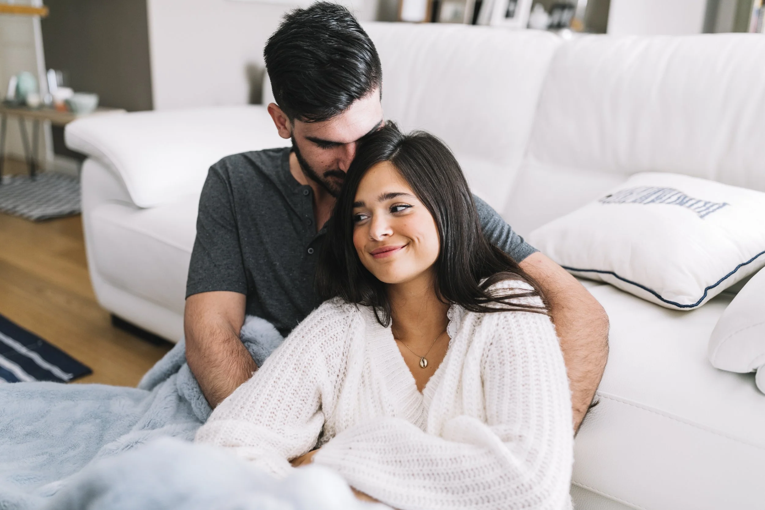 A happy couple sitting on the floor in a cozy living room, the man with dark hair and beard kissing the woman's forehead, she has long dark hair and is smiling with her eyes closed, wearing a white knit sweater.