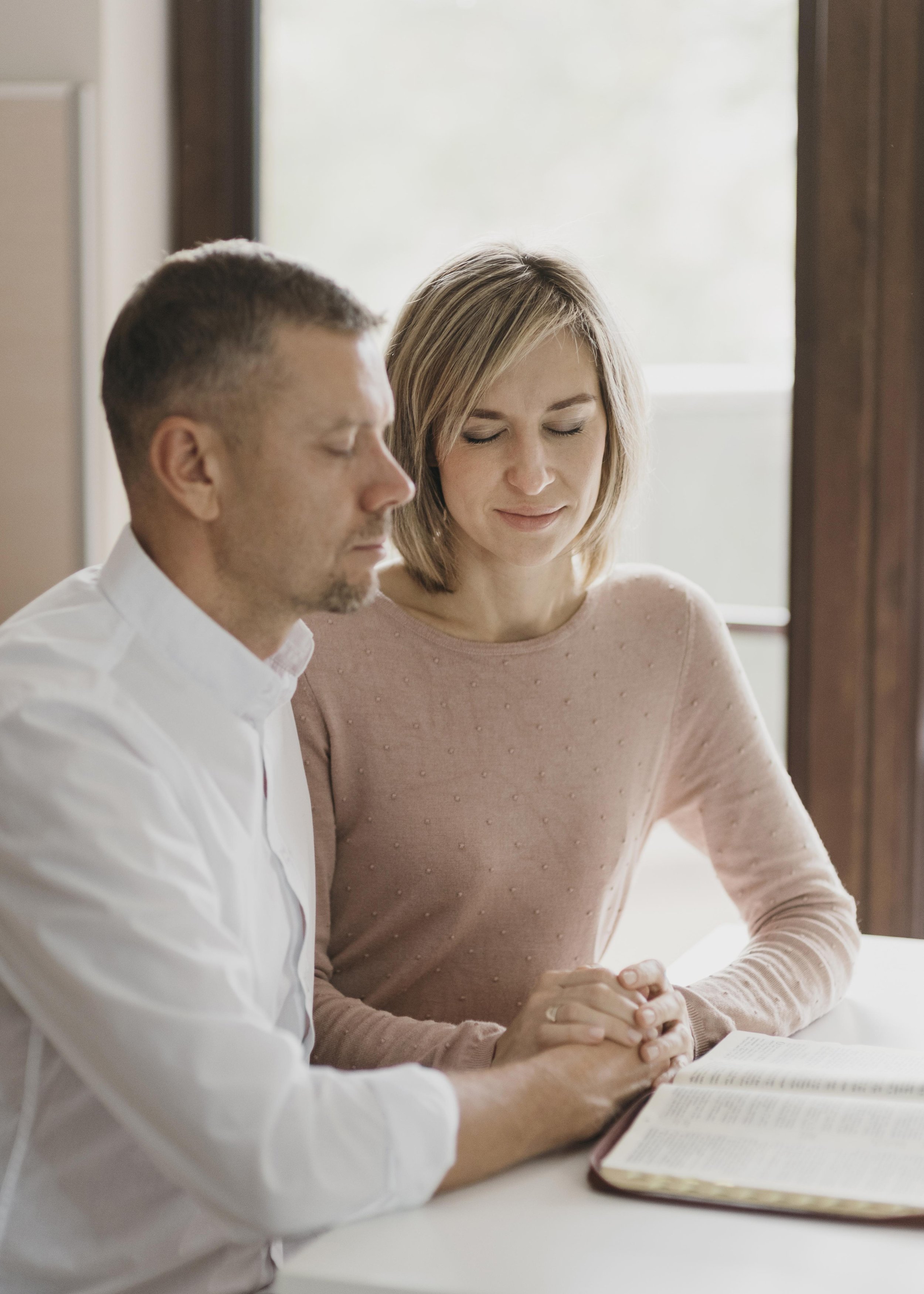A man and a woman sitting together at a table with a book, in a room with natural light coming through a window, both with eyes closed, hands clasped, in a moment of prayer or reflection.