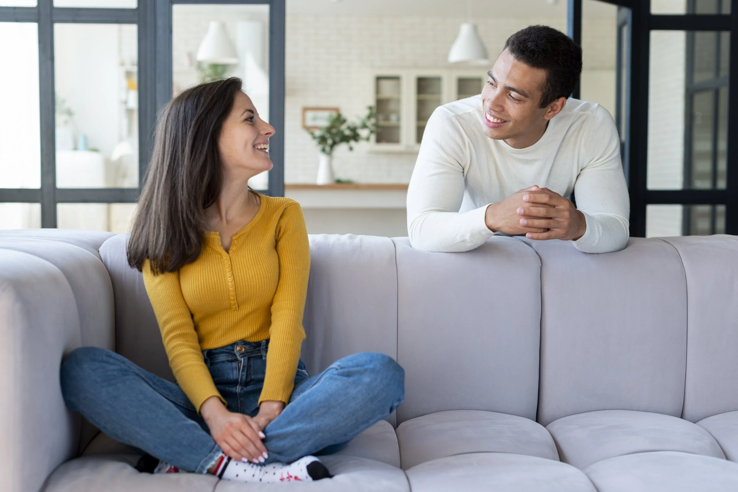 A woman and a man are sitting on a sofa in a bright, modern home, smiling at each other.