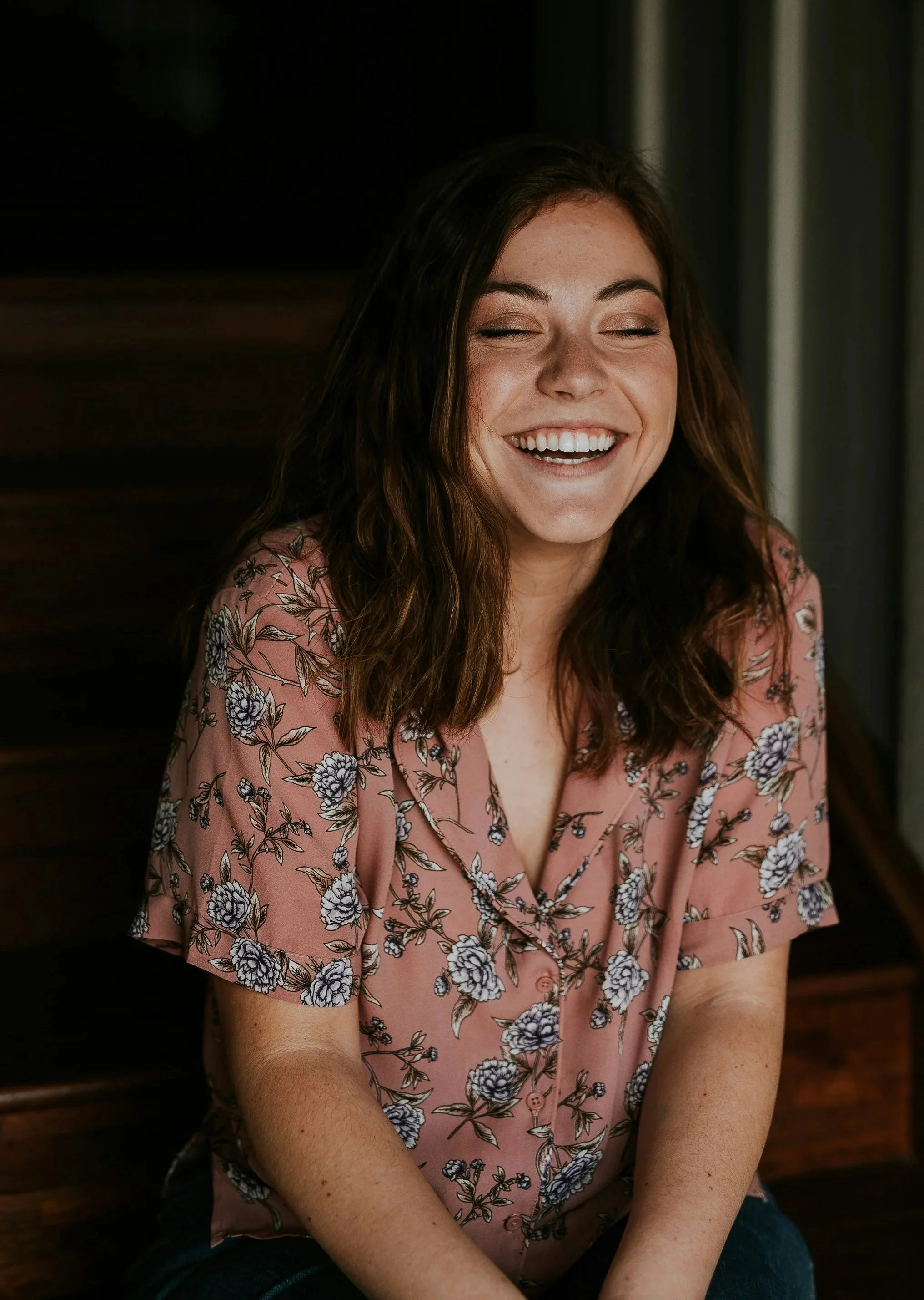 A woman with brown hair smiling with eyes closed, wearing a pink floral blouse, sitting against a dark background.