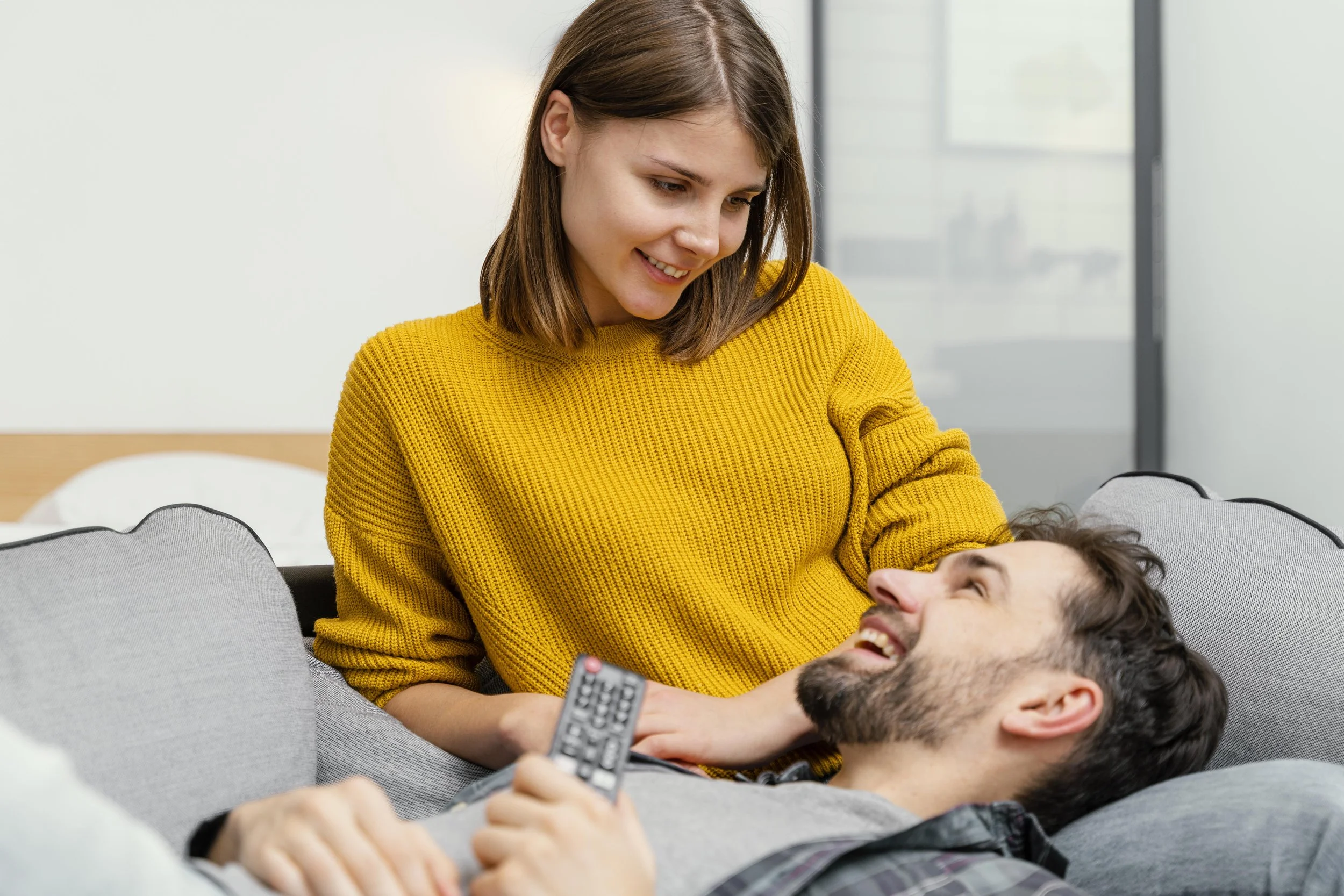 A woman in a yellow sweater is smiling and looking down at a man lying on a couch, who is holding a TV remote and smiling back at her.