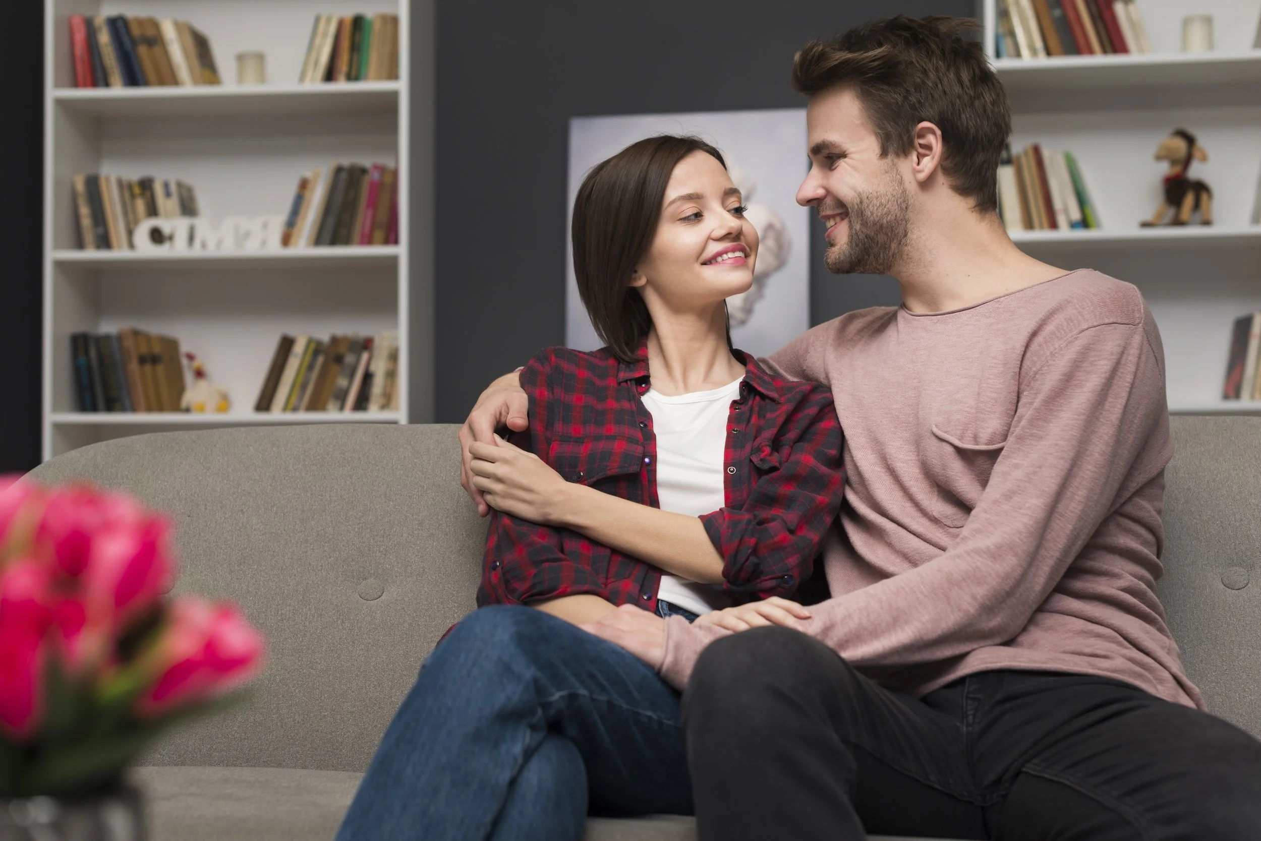 A young couple smiling and looking into each other's eyes while sitting on a couch. The woman is wearing a red plaid shirt, and the man is wearing a light pink shirt. They are in a living room with bookshelves in the background.
