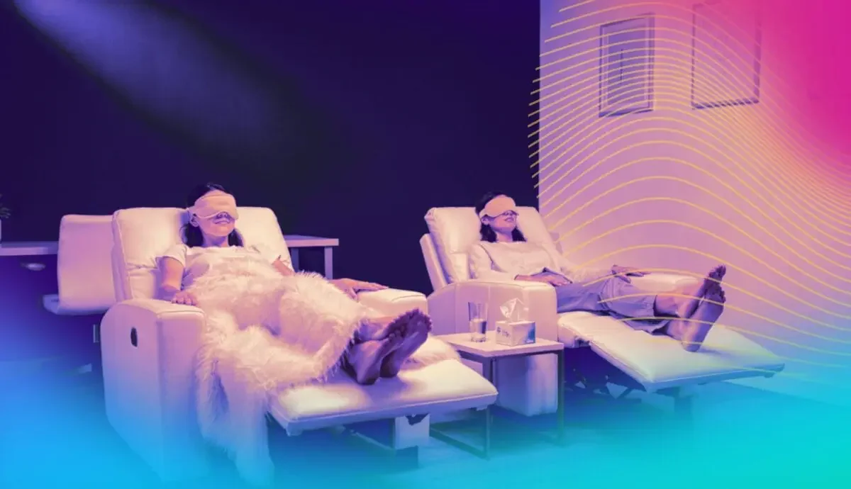 Two women relaxing in recliner chairs with blindfolds on, in a room with colorful lighting and a digital wave pattern projected on the wall.
