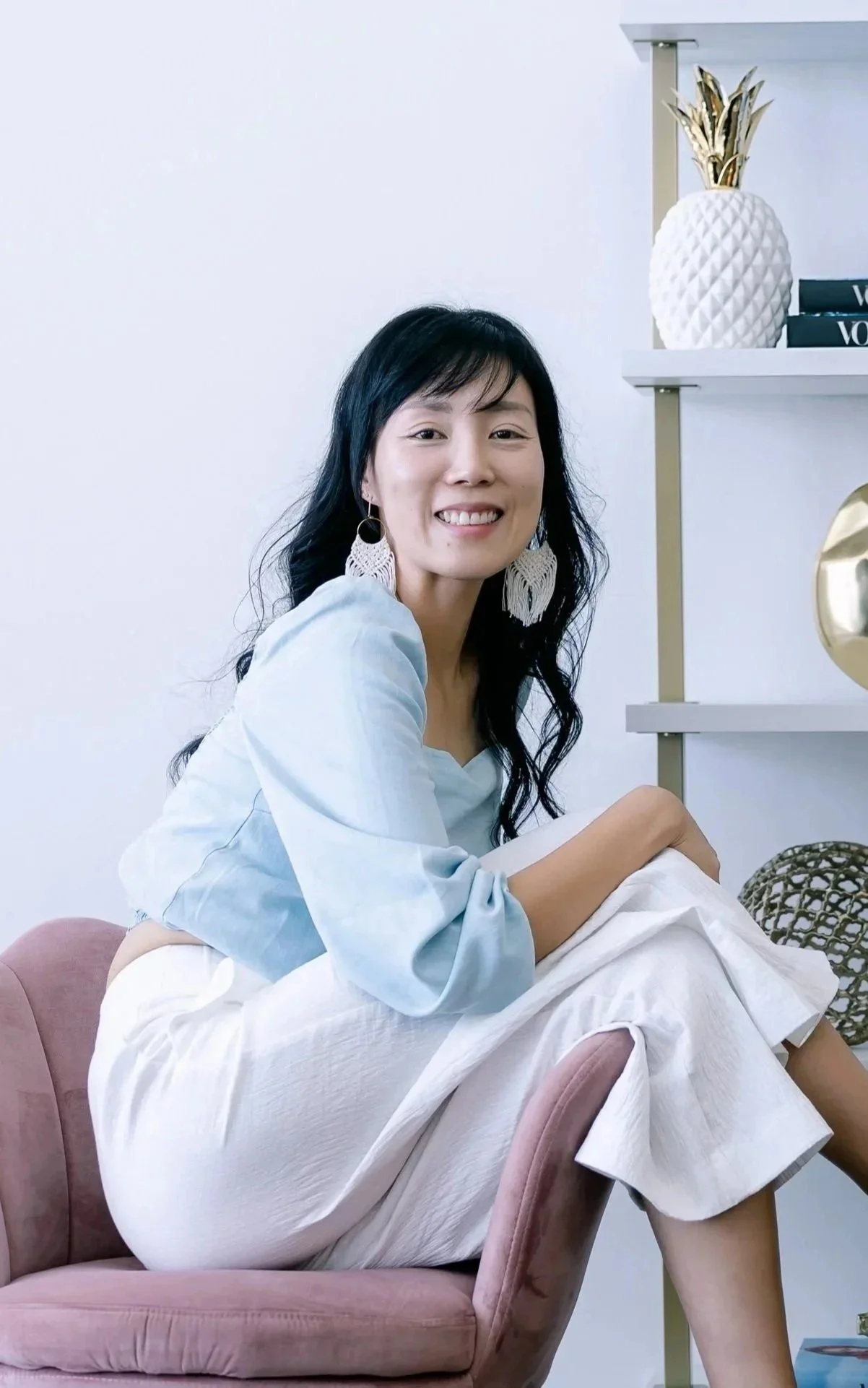 A woman with dark hair, wearing large white earrings and a light blue top, sits on a pink velvet armchair in a room with white walls, smiling at the camera. Behind her are bookshelf shelves with decor including a white pineapple-shaped vase and books.