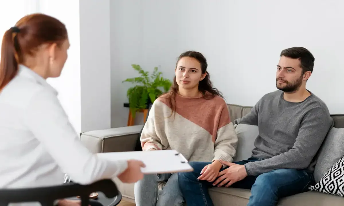 A woman and man sitting on a couch during a therapy session with a therapist taking notes.