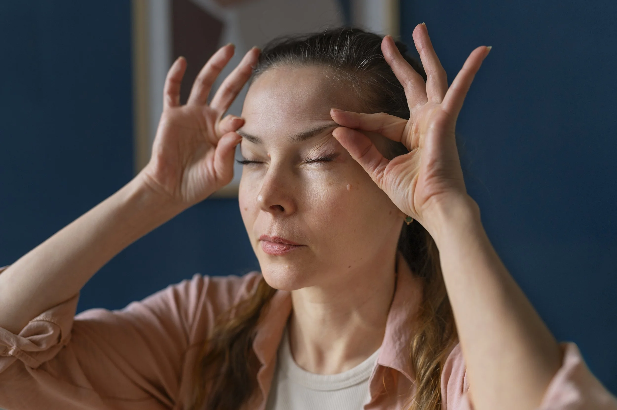A woman with brown hair and closed eyes is performing a facial massage or relaxation technique, with her fingers gently pressing on her forehead and around her temples.