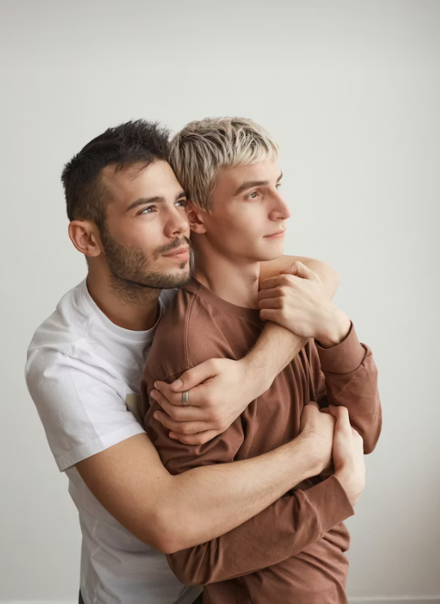 Two men hugging each other, one embracing the other from behind, both looking to the right. They are standing against a plain, light-colored background.