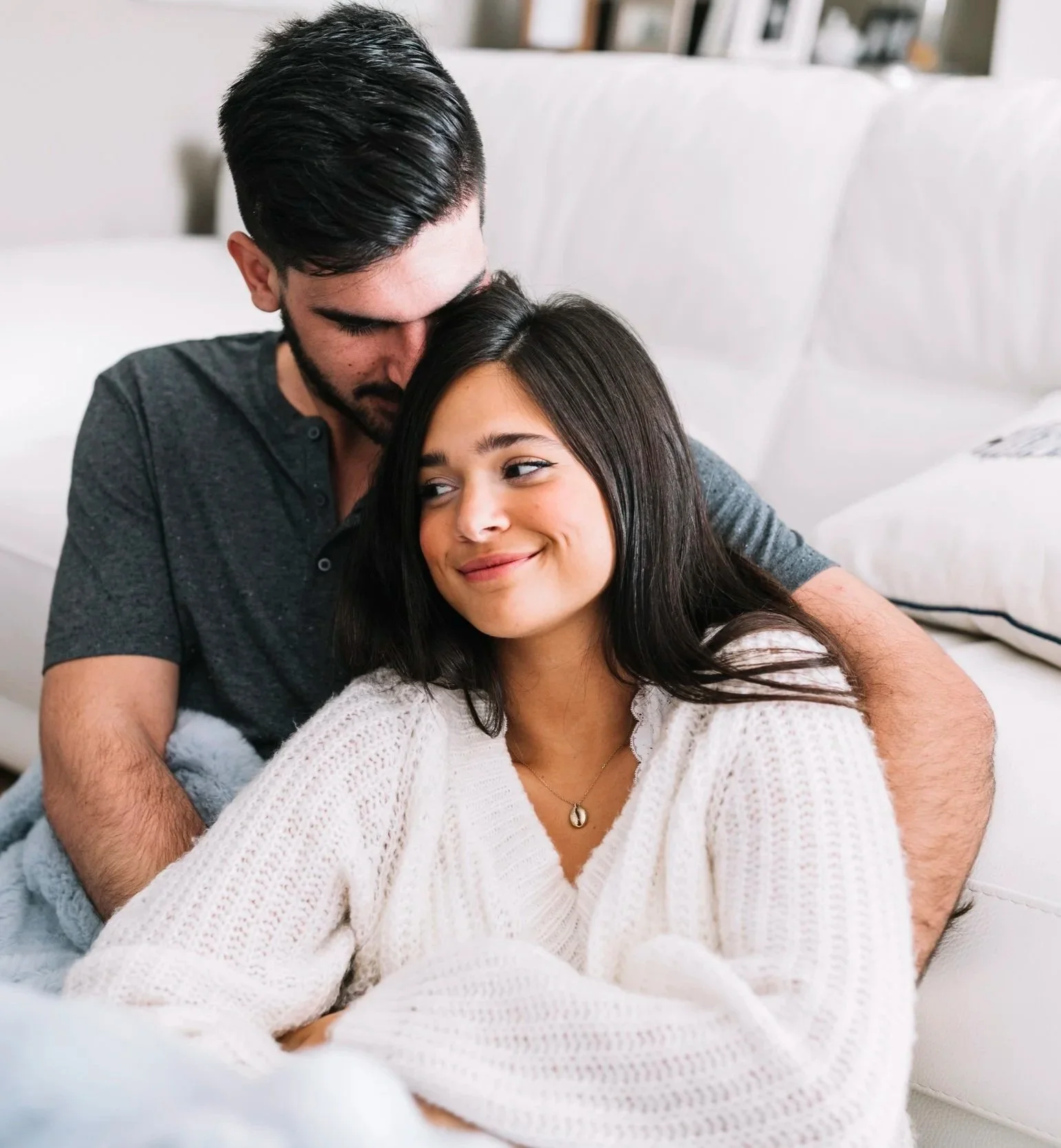 A young man and woman sitting close together on a white couch in a living room, with the man resting his head on the woman's forehead and both smiling softly.