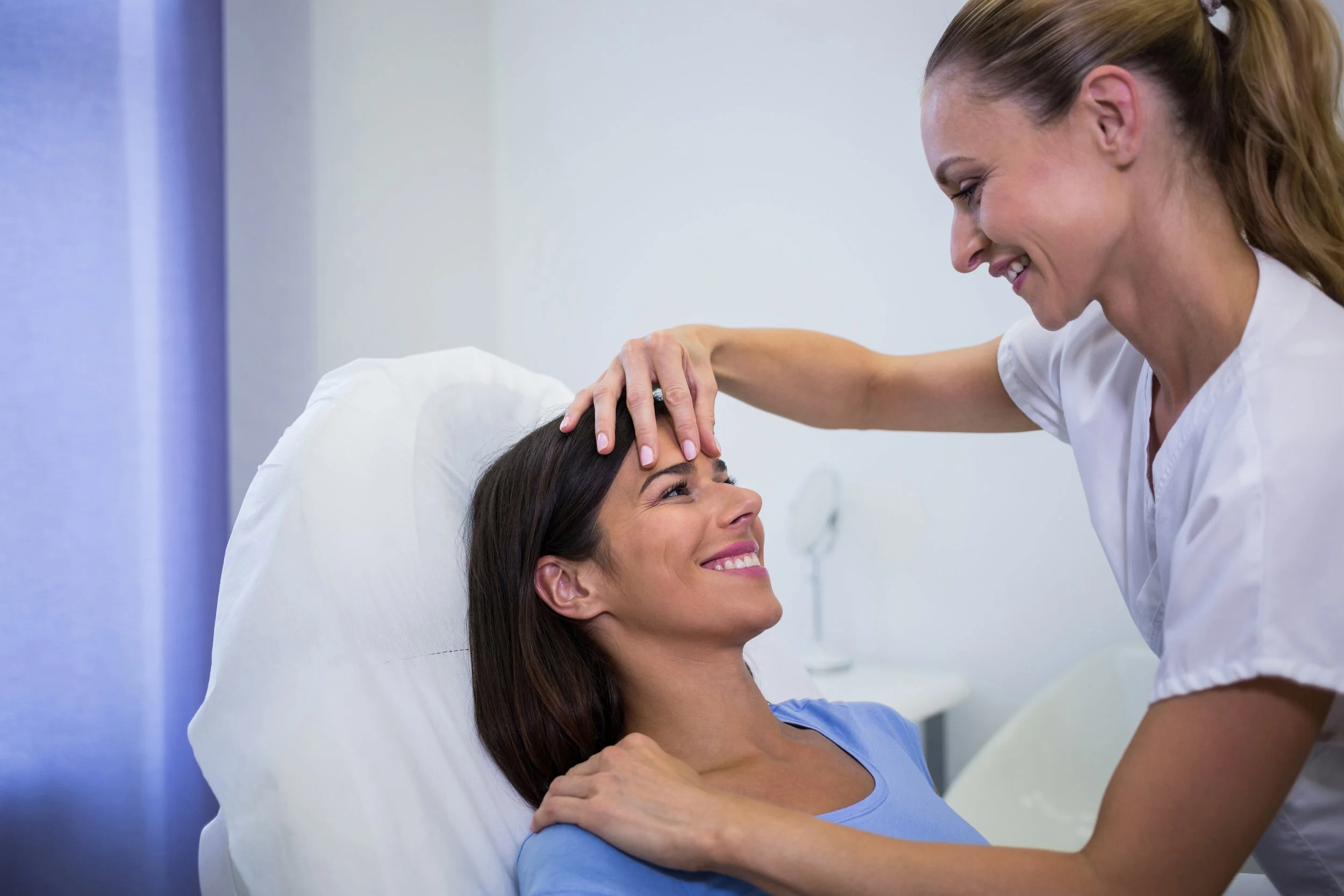 A woman lying in a hospital bed smiling while a nurse or doctor gently touches her forehead. The healthcare professional is smiling, and the woman appears happy and comfortable.