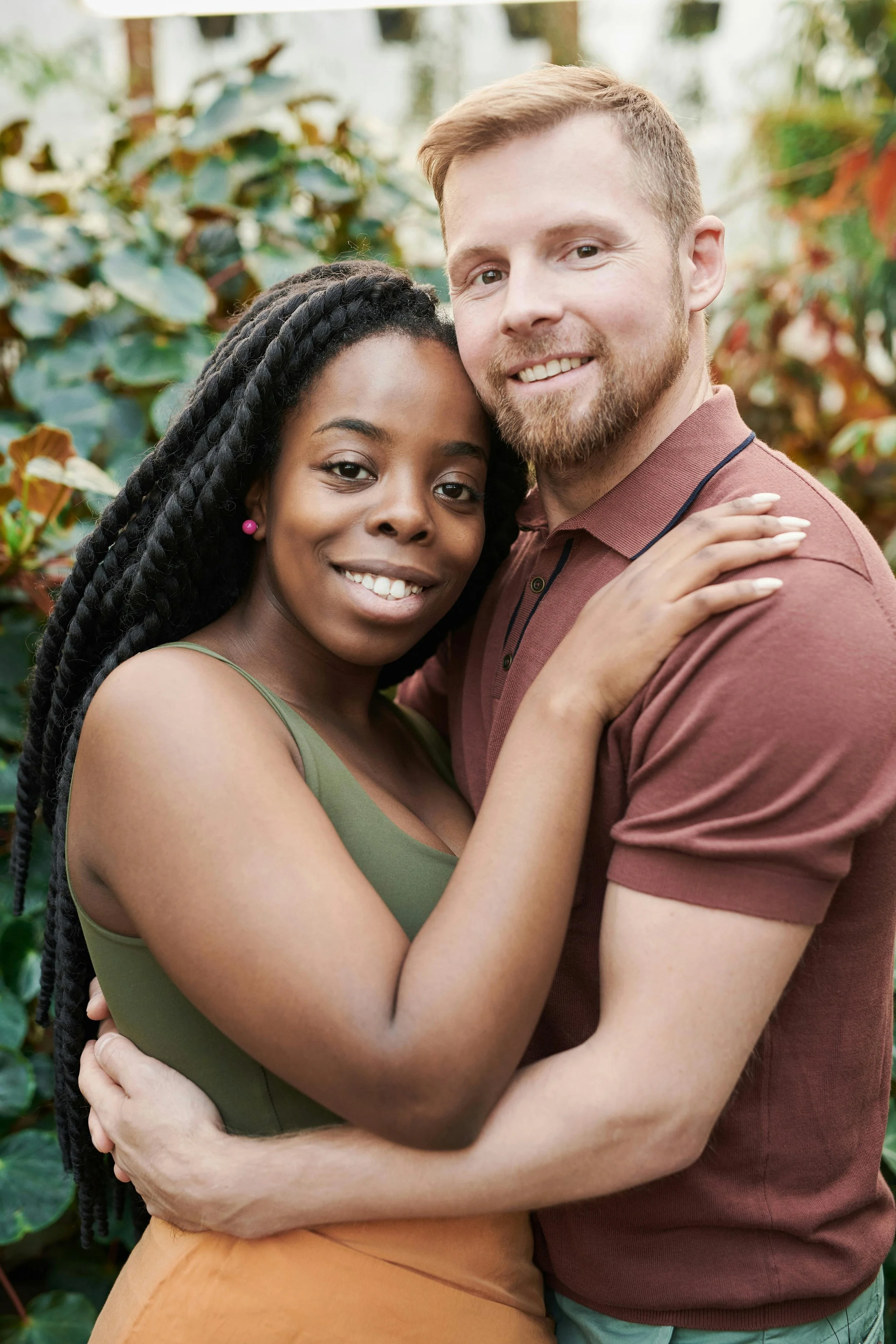 A woman with long braided hair and earrings hugging a man with a beard and short hair, both smiling, standing outdoors with green foliage in the background.