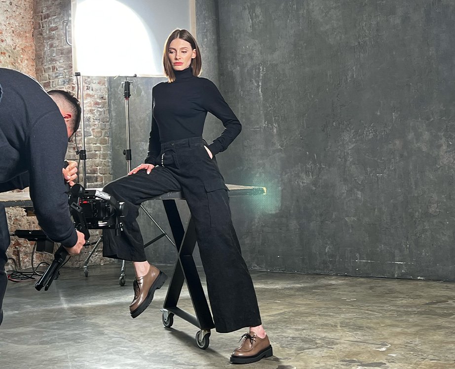 Fashion photoshoot in industrial setting with woman posing on stool and cameraman adjusting equipment.