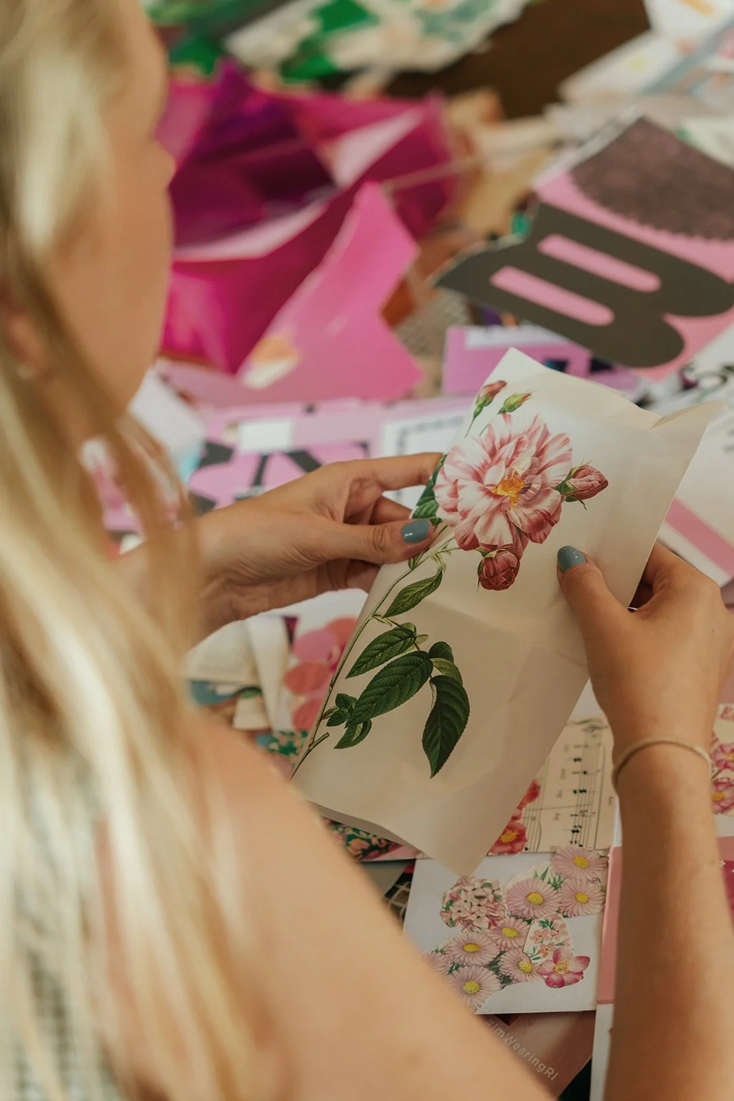 A woman holding a botanical print of a pink flower with green leaves, surrounded by various colorful papers and craft supplies on a table.