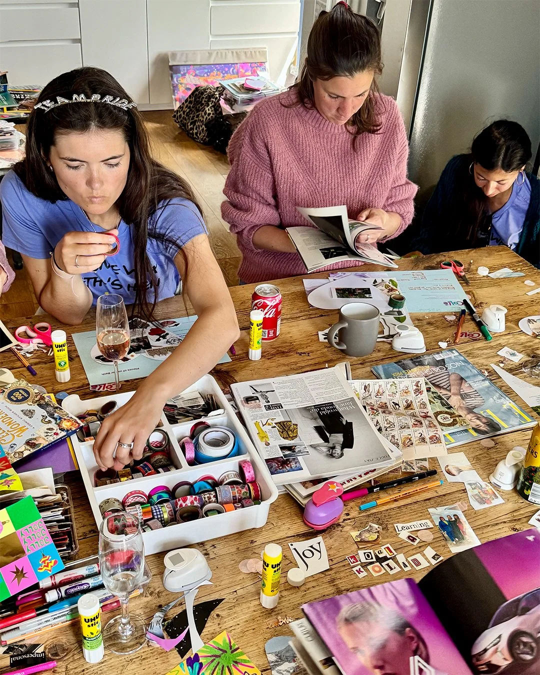 A group of women working on a craft project at a wooden table. The table is cluttered with colorful washi tapes, magazines, glue sticks, scissors, markers, and assorted craft supplies. One woman is handling washi tape, another is looking through a magazine, and the third woman is focusing on her craft tasks.