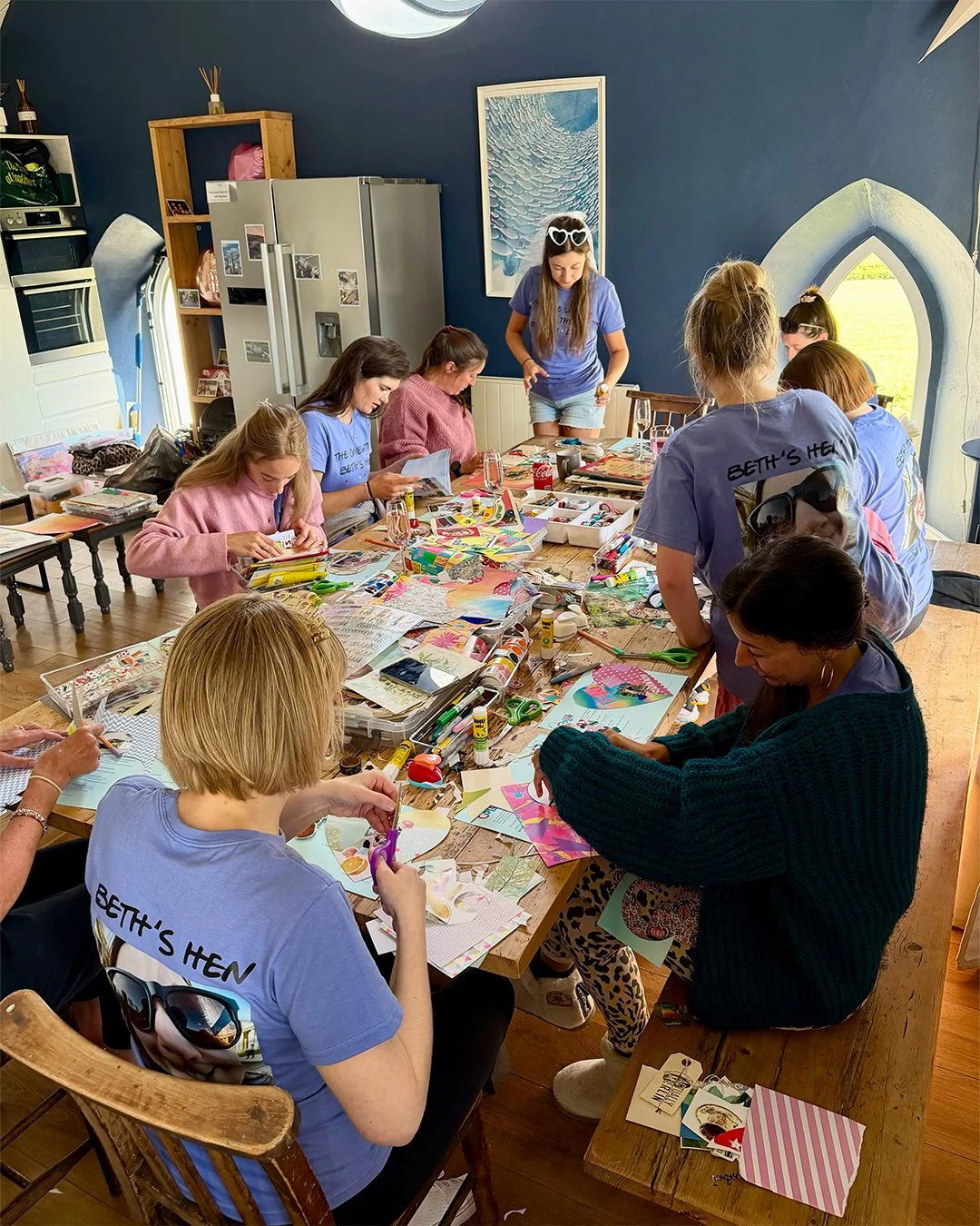 Multiple people are gathered around a large table engaged in arts and crafts projects, with various supplies scattered on the table, in a cozy room with blue walls and a window with a decorative arch.