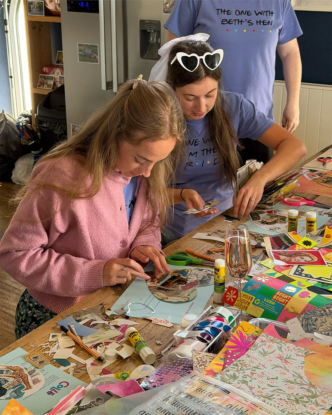 Two women are engaged in a craft project at a table, surrounded by colorful paper, scissors, glue, and other craft supplies, while another person stands in the background.