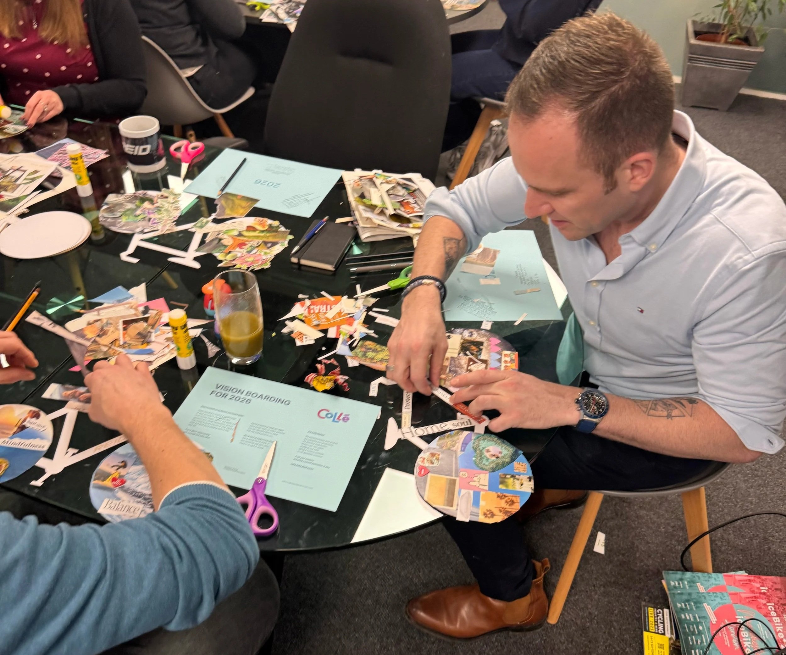 People sitting at a table working on a craft project with paper, scissors, glue, and magazines.