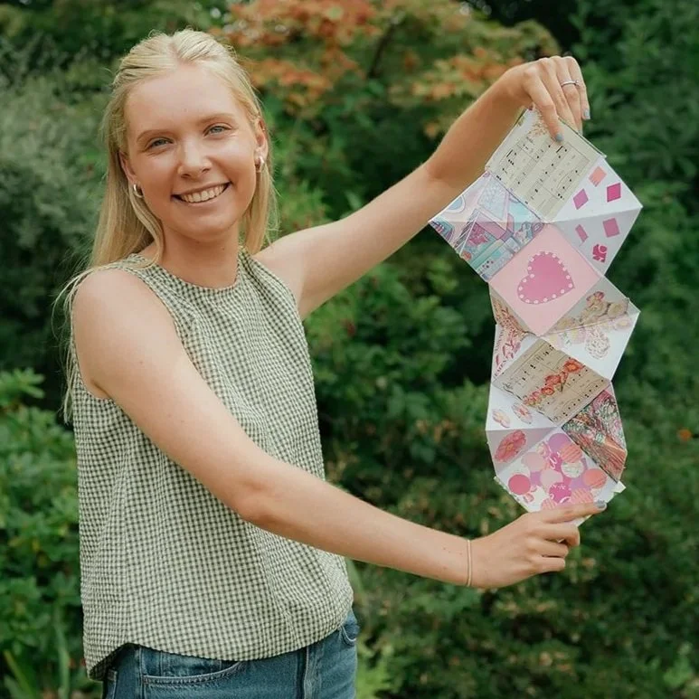 A smiling young woman with blonde hair in a checkered sleeveless top and jeans holds a chain of colorful paper crafts outdoors with greenery in the background.
