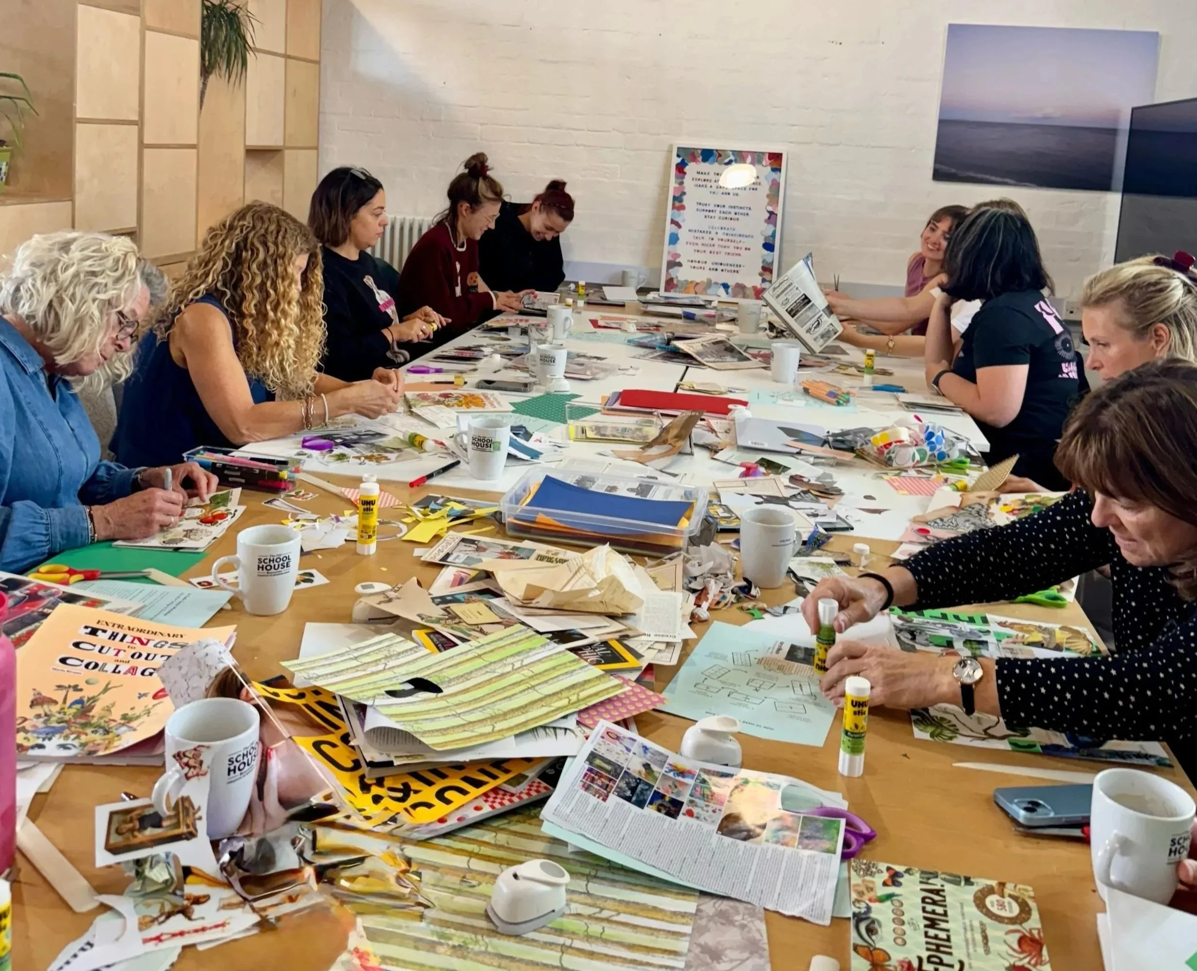 A group of people sitting around a long table engaged in arts and crafts activities, with various papers, magazines, scissors, glue sticks, and coffee mugs scattered across the table.
