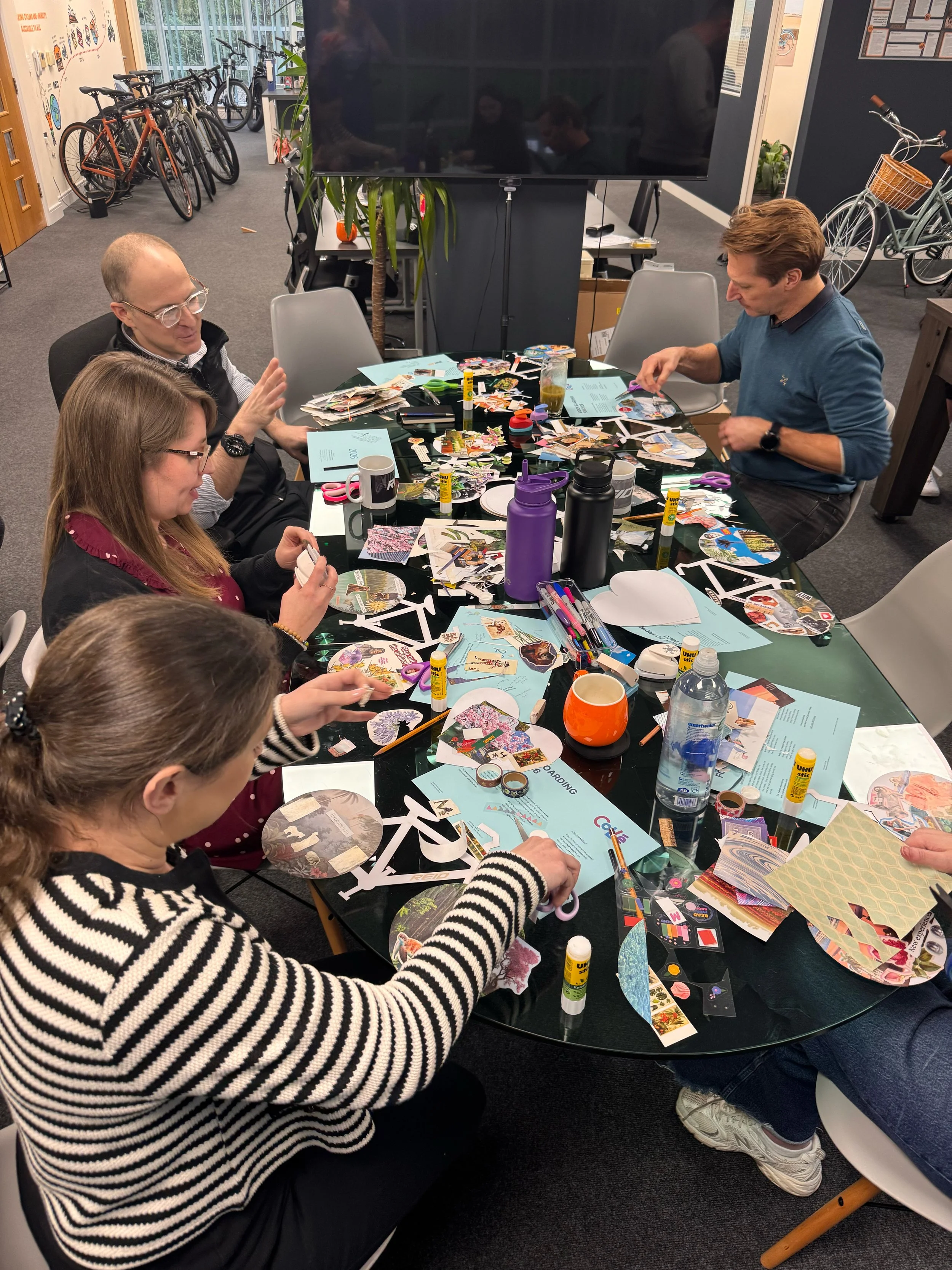 People gathered around a table creating and decorating paper crafts, with various scissors, glue sticks, and decorative paper scattered across the table. Bicycles and a large screen are visible in the background.