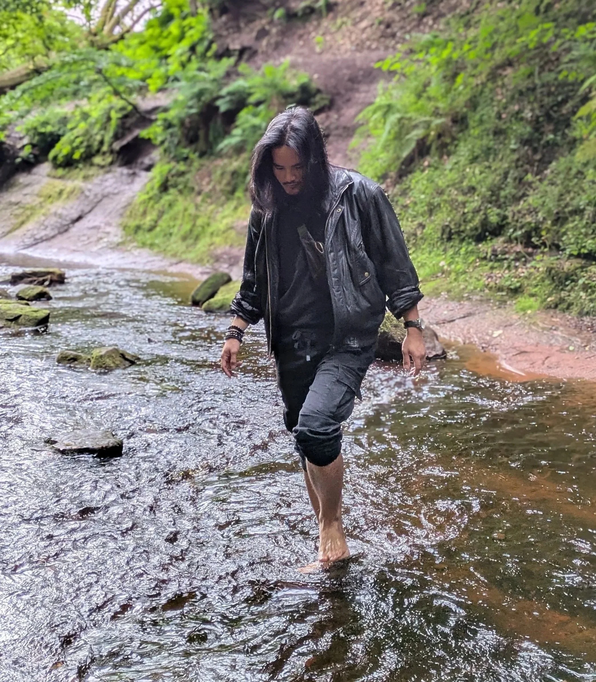 A person with long black hair wearing a black leather jacket, black t-shirt, and rolled-up pants stands ankle-deep in a shallow stream in a lush, green forest.