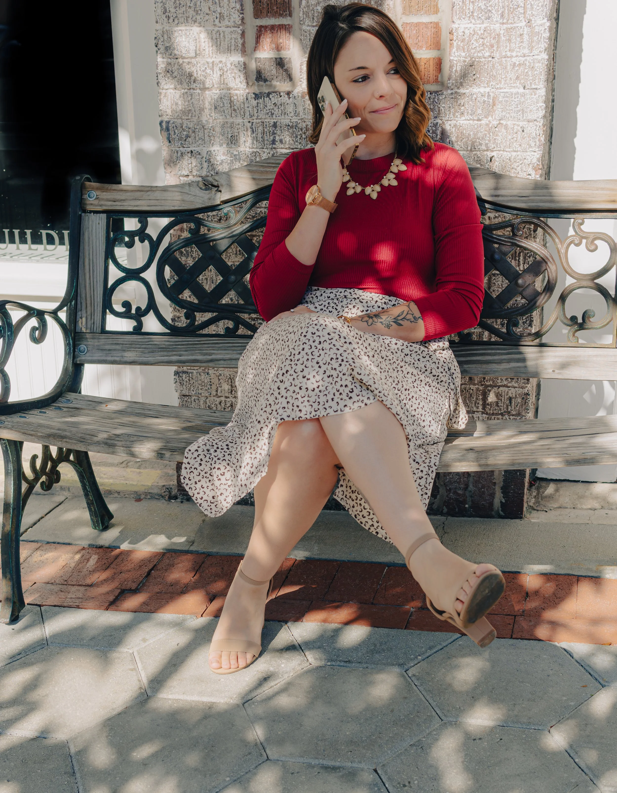 A woman wearing a red top and a leopard print skirt, sitting on a wooden and metal bench outside while talking on her smartphone.