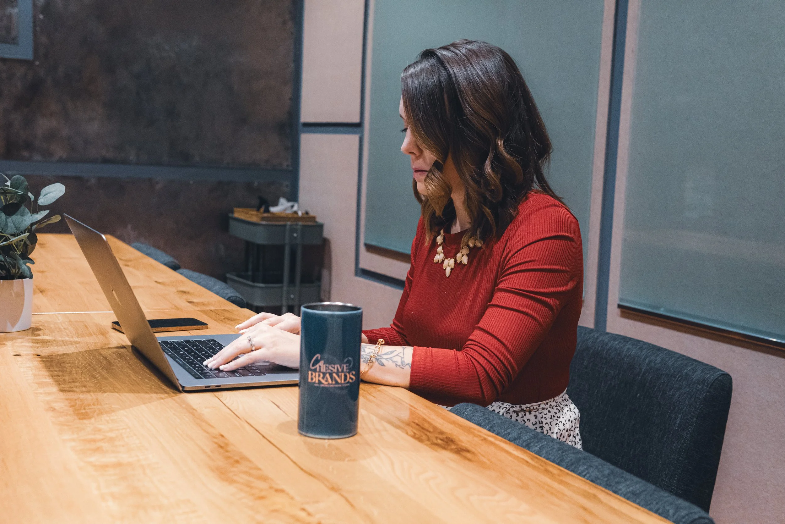 A woman with shoulder-length dark hair, wearing a red top and a statement necklace, works on a laptop at a wooden table in a modern office or conference room. A blue mug with the words "Creative Brands" is in front of her.